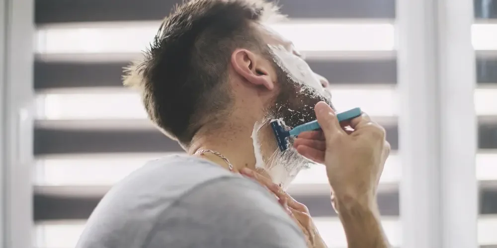 Man shaving his bearded jaw with an electric razor and shaving cream in a bright bathroom, creating a calm, everyday grooming scene.