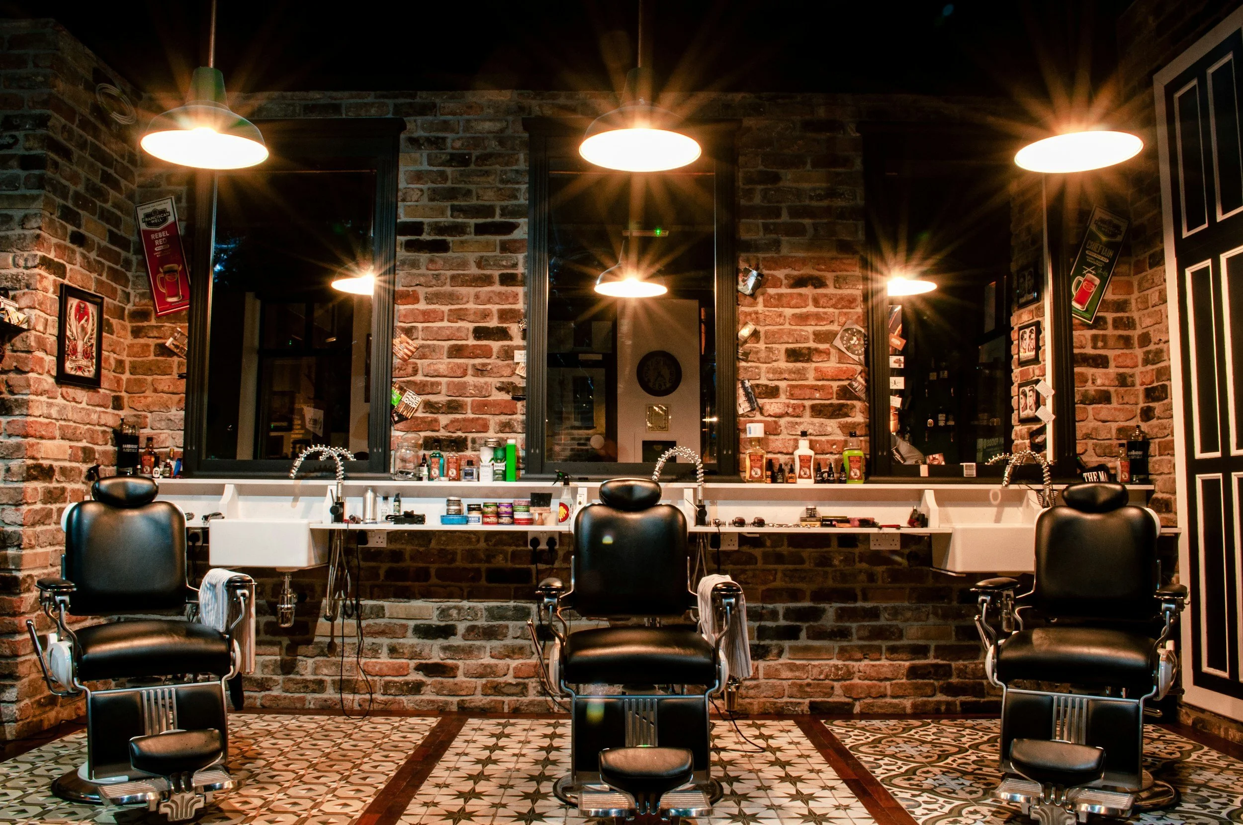 Vintage barbershop with three black chairs, illuminated by warm lighting. Exposed brick walls and ornate floor tiles create a nostalgic ambiance.