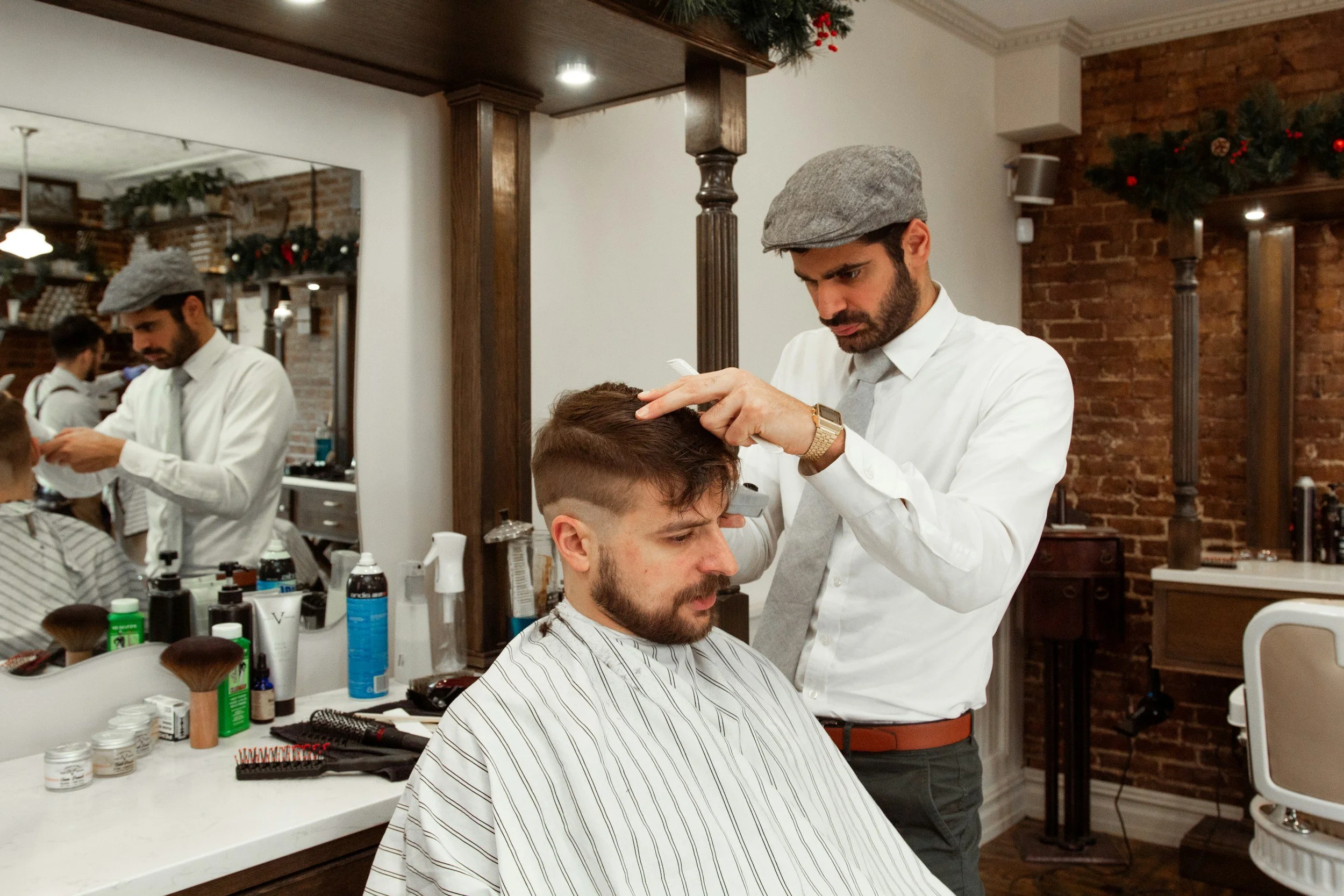 Barber in a white shirt and flat cap trims a man's hair in a vintage-style barbershop with brick walls. Reflective mirrors and hair products visible.