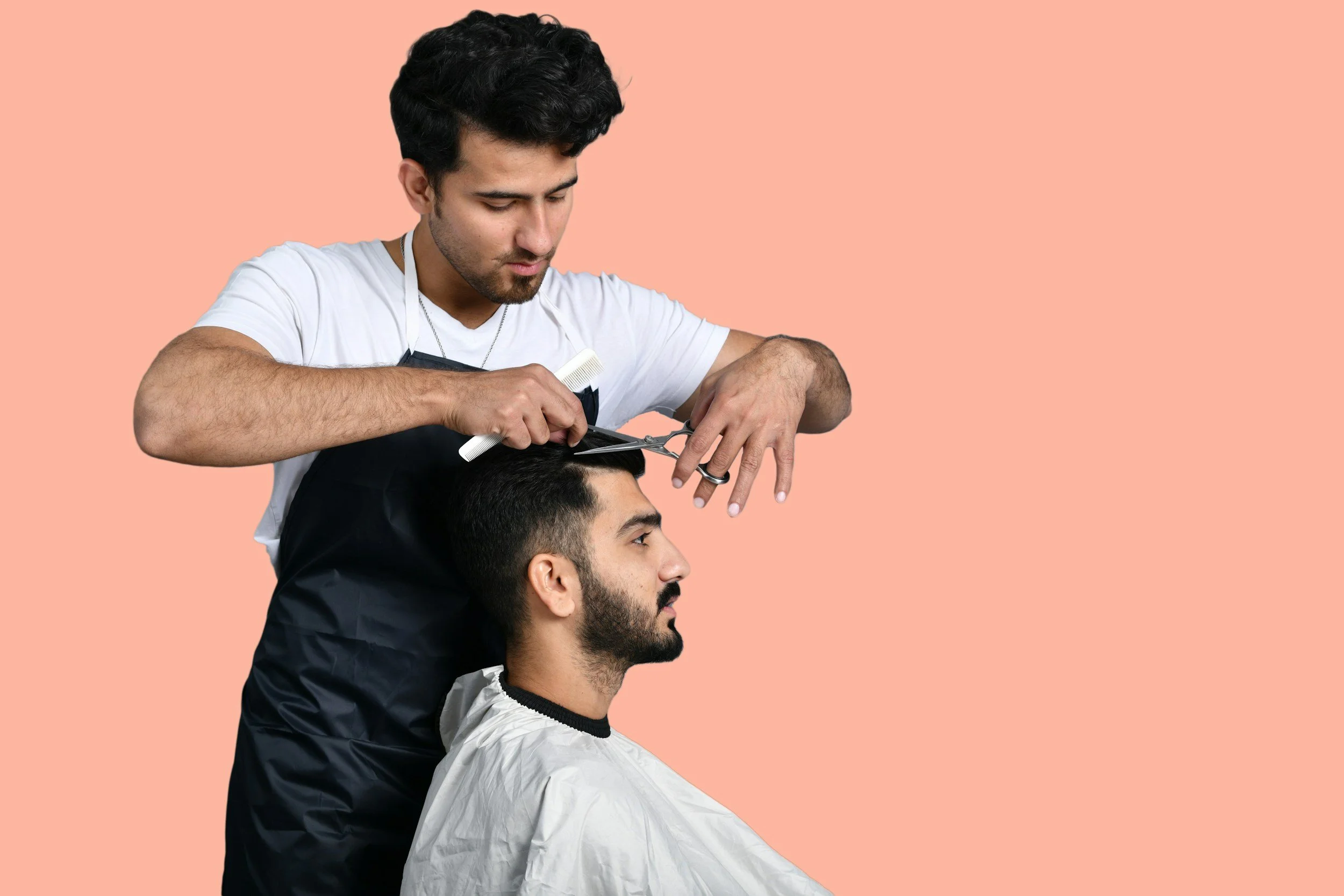 A barber in a white shirt and black apron cuts a seated customer's hair against a peach background. The scene conveys focus and professionalism.