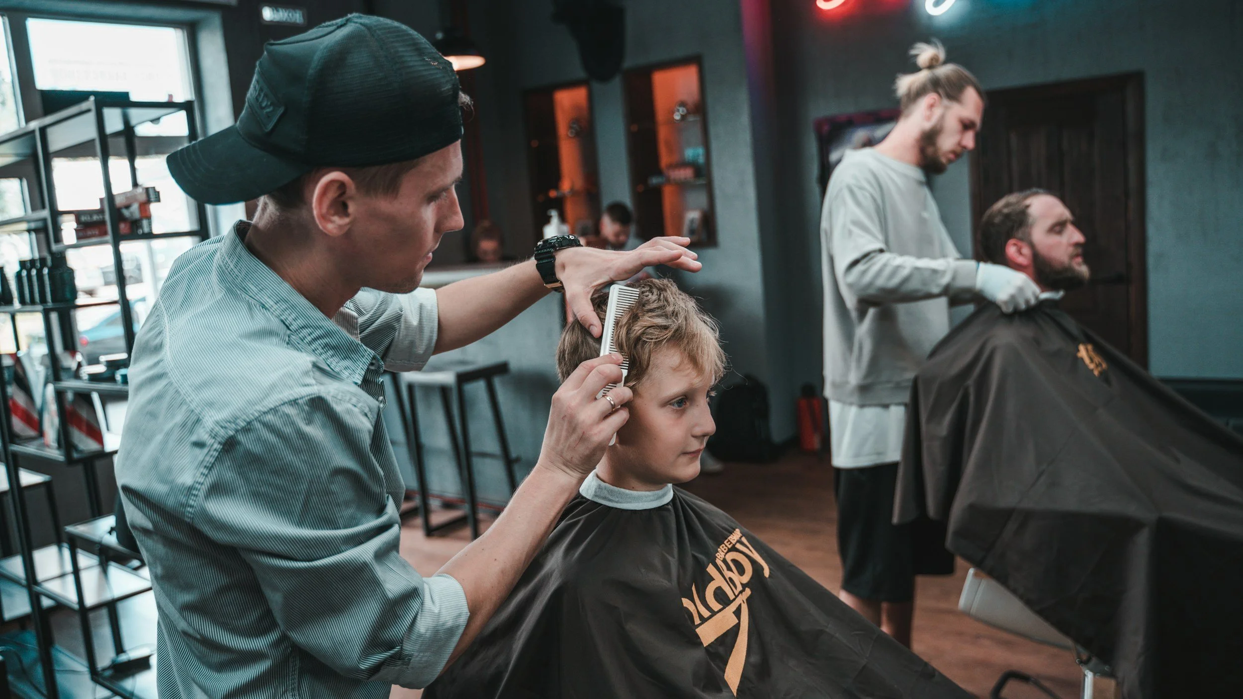 A modern barbershop with two barbers styling clients' hair. The atmosphere is relaxed, featuring dim lighting and wooden floors, conveying a trendy vibe.