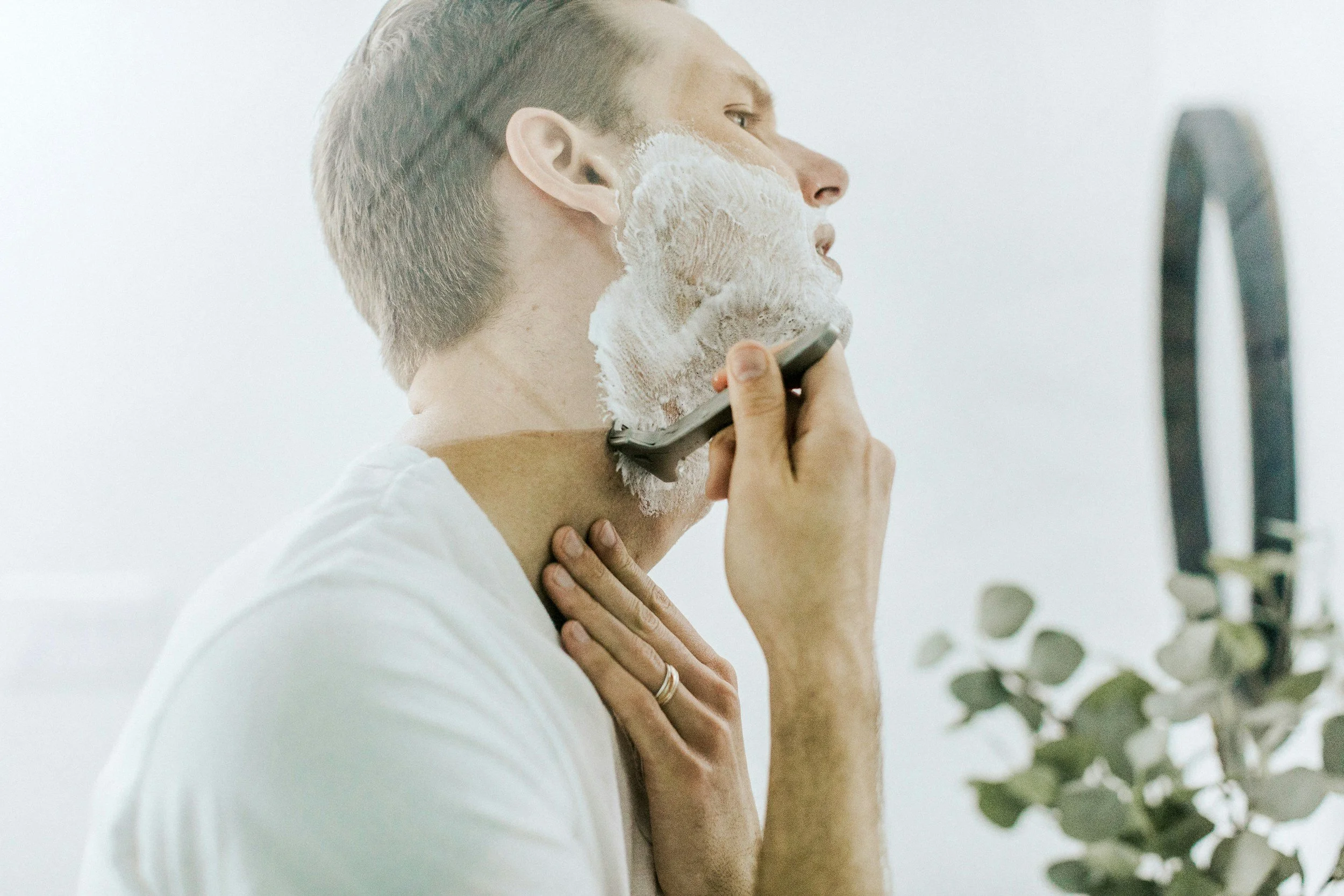 A man applies shaving cream to his face while looking in a bathroom mirror. He wears a white shirt, and plants are visible in the background.