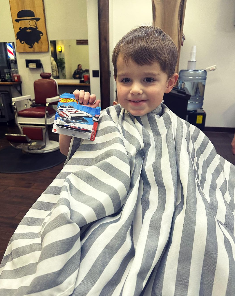 A young boy with brown hair is sitting in a barber's chair wearing a gray and white striped barber cape, holding a Hot Wheels toy car package, inside a barber shop.