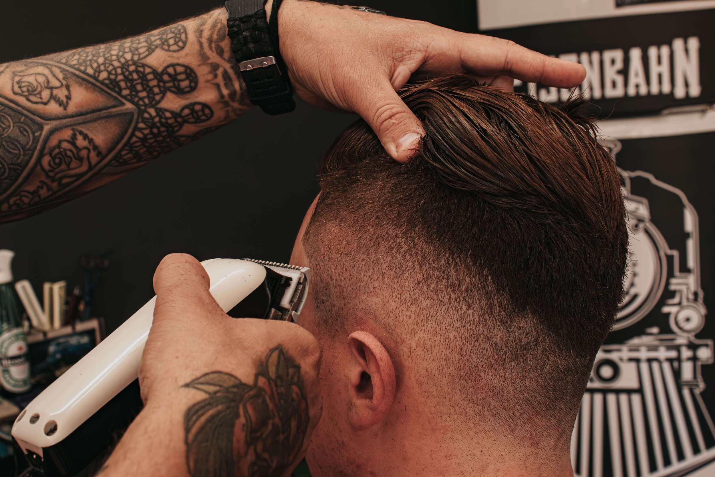 Tattooed barber expertly trims a client's brown hair with clippers, creating a fade haircut. The scene conveys focus and precision.