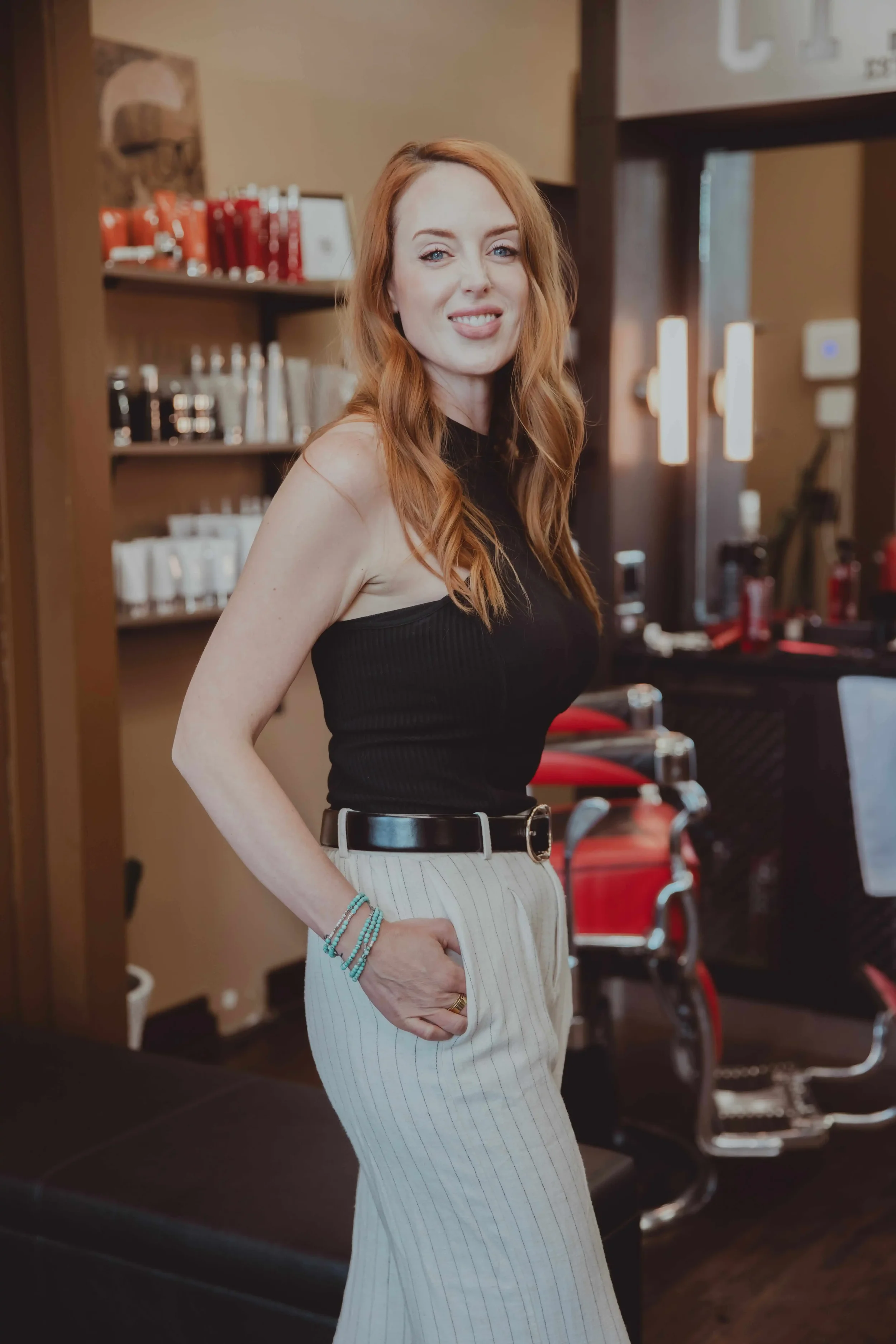A woman with long red hair, wearing a black sleeveless top and white striped pants, standing indoors with her hand on her hip and smiling. Background shows shelves with various bottles and a red wheelchair.