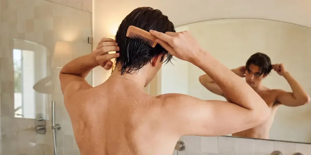 A shirtless man combs his wet dark hair while looking in a bathroom mirror, water droplets on his back in a calm post-shower grooming moment.