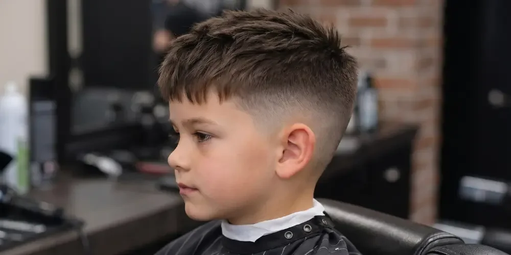 Young boy in profile sits calmly in a barbershop chair, wearing a black cape and showing a fresh textured fade haircut near a brick wall.