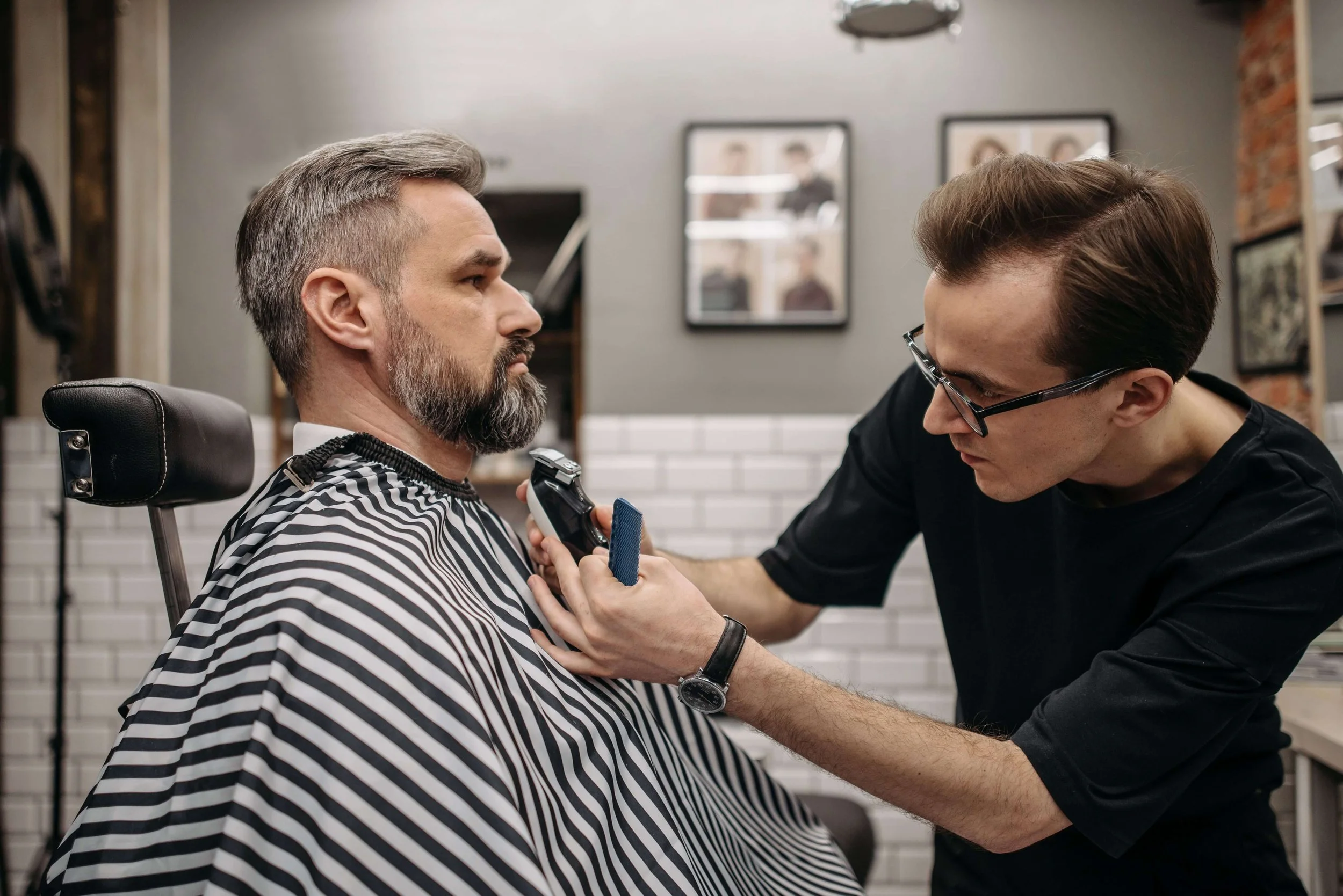 A barber wearing glasses carefully trimming a middle-aged man's beard with hair clippers in a barbershop.