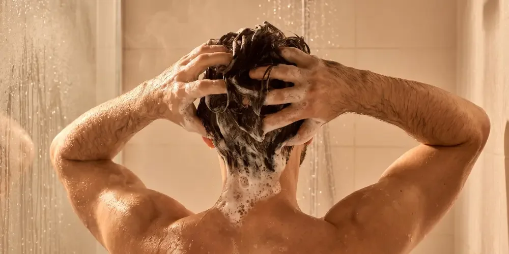 A person massages shampoo into wet hair beneath a shower, arms raised as water runs down; warm beige tiles create a calm, private mood.