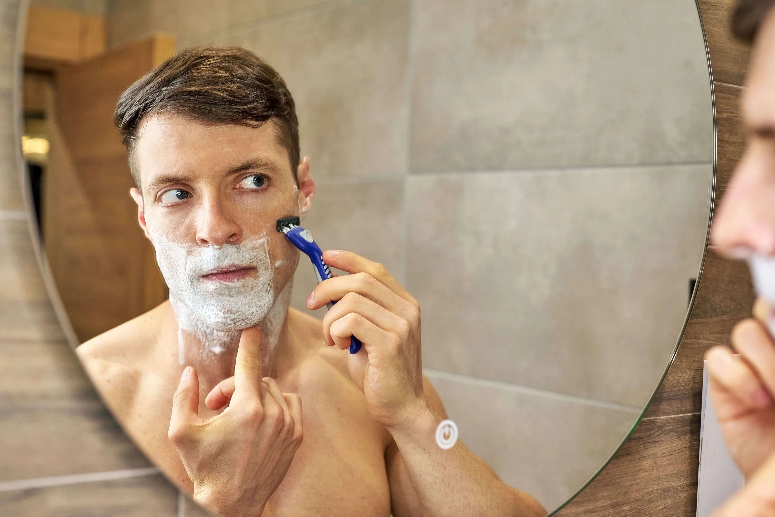 Man shaving in front of a bathroom mirror, applying razor to foam-covered face. He looks concentrated, with neutral-toned tiles in the background.