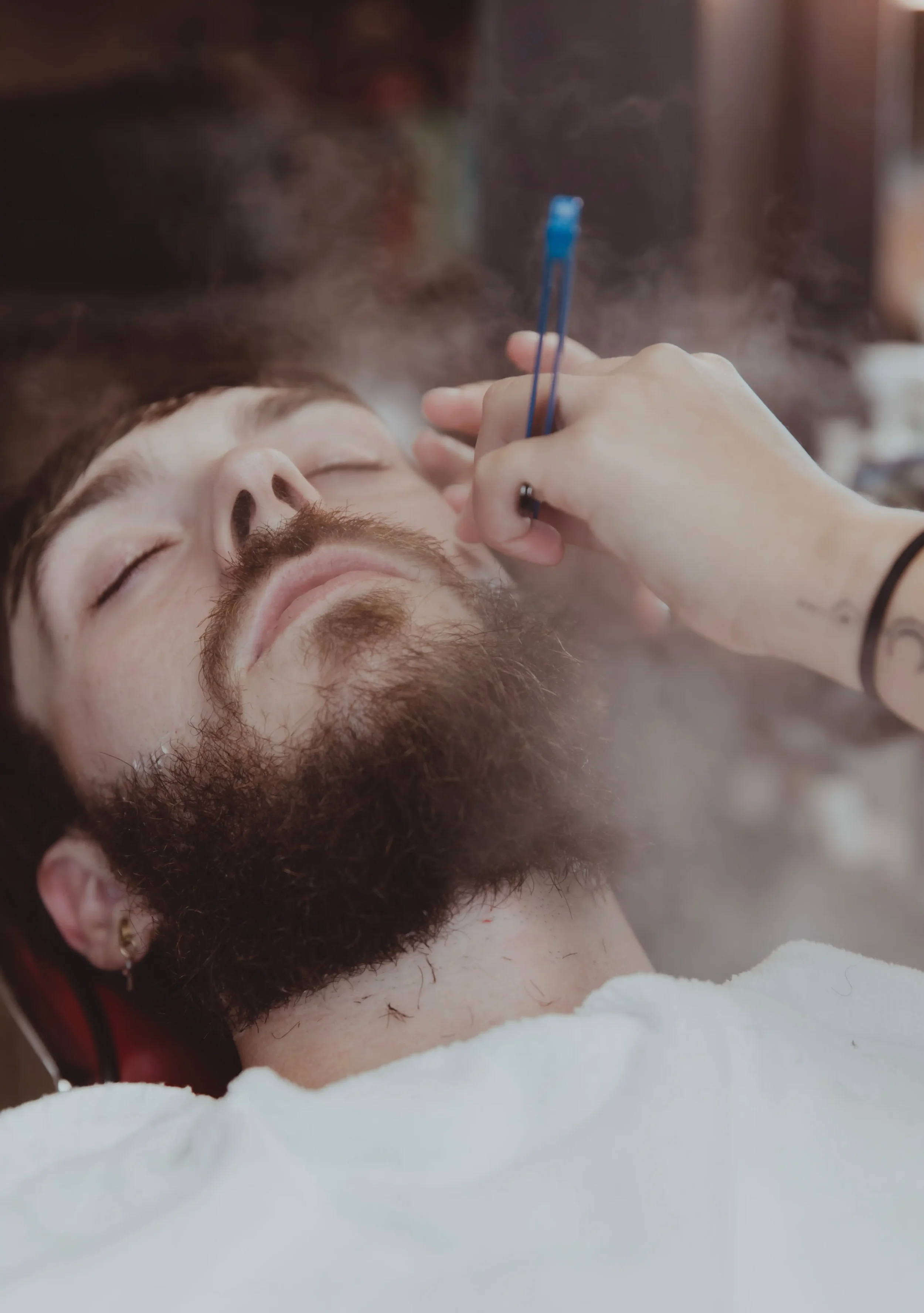 A man with a beard and closed eyes lying down, getting a facial treatment with a tool held by a person with a bracelet, and steam around his face.