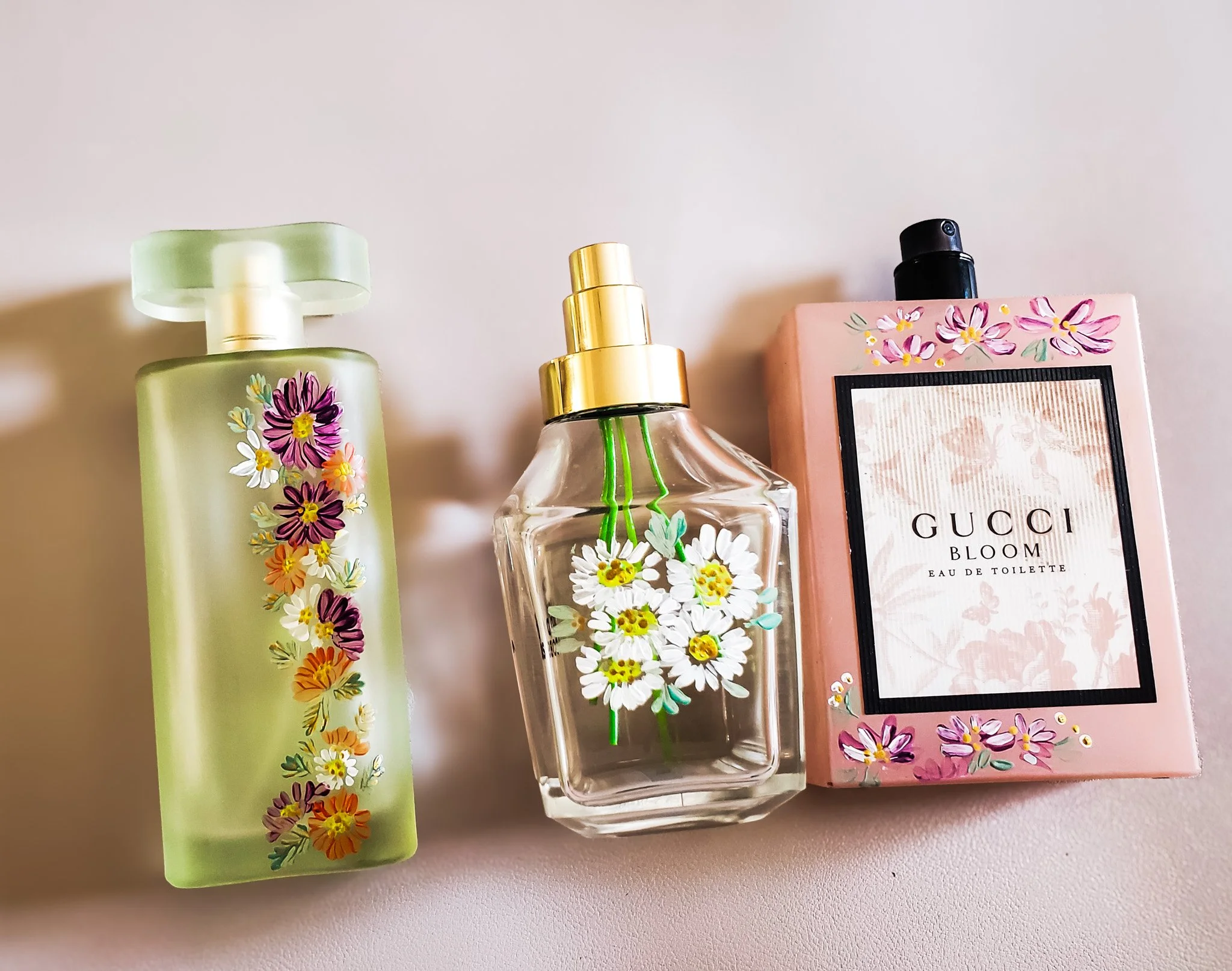 Three floral-scented perfume bottles, one with a green frosted top and floral design, a clear glass bottle with daisy flowers, and a pink Gucci Bloom Eau de Toilette with floral design.
