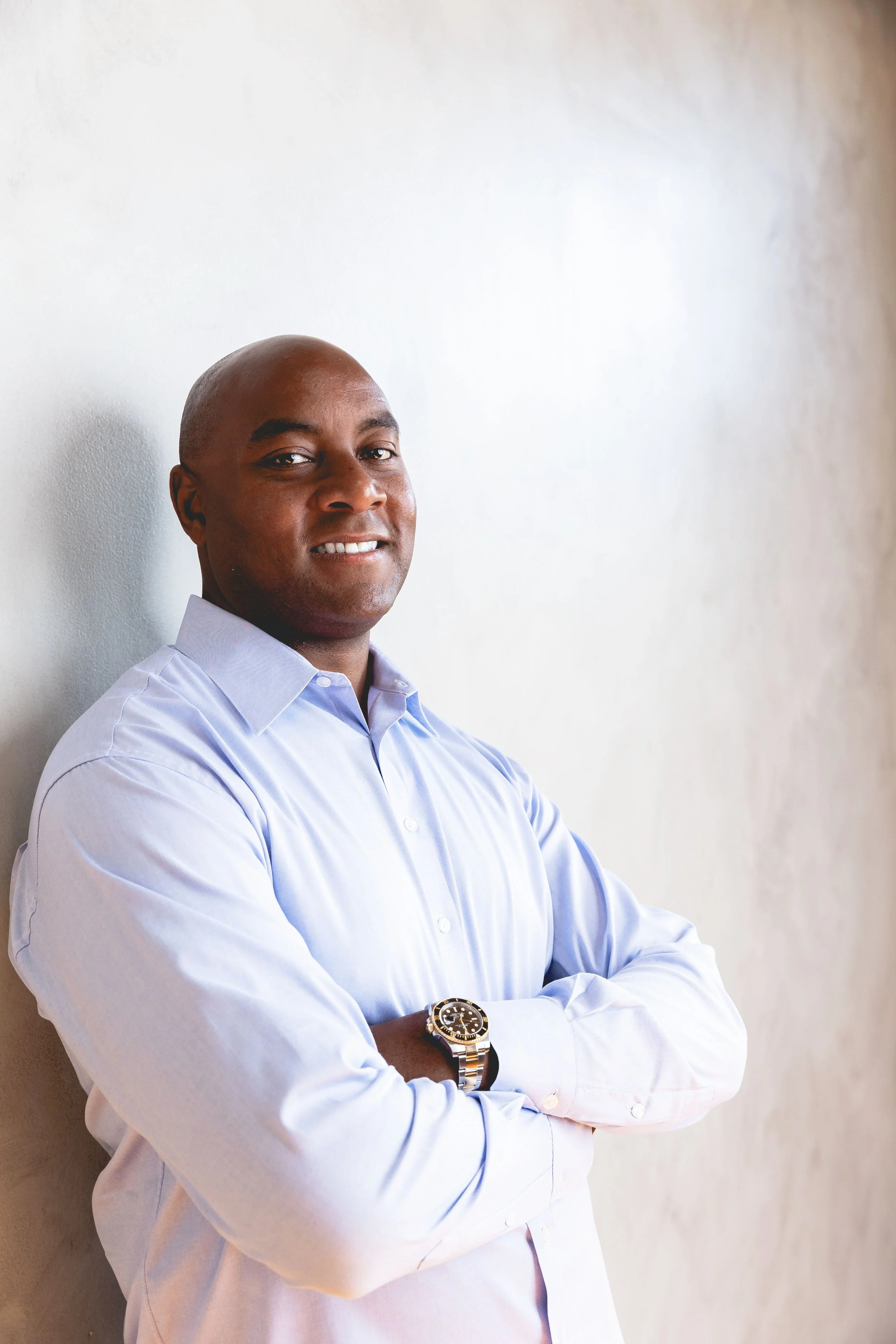 Mario Hambrick, a professional man with dark skin, dressed in a black suit, white shirt, and purple patterned tie, sitting by a large window with a clean background view, smiling at the camera.