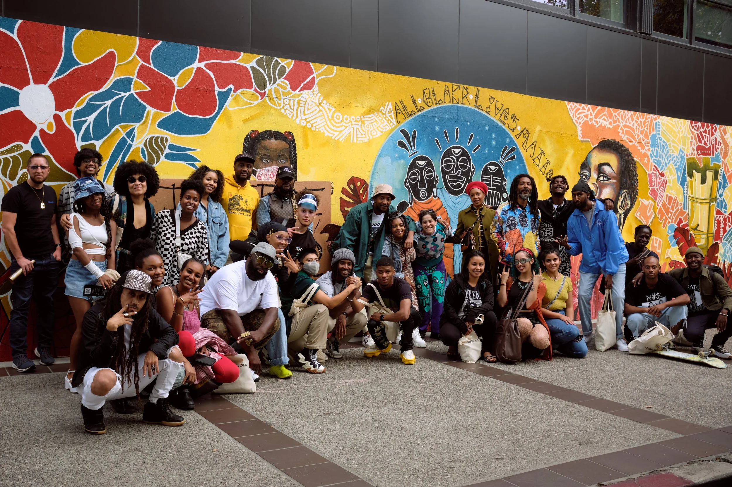 A diverse group of people posing in front of a colorful mural featuring faces, flowers, and abstract designs on an outdoor wall.