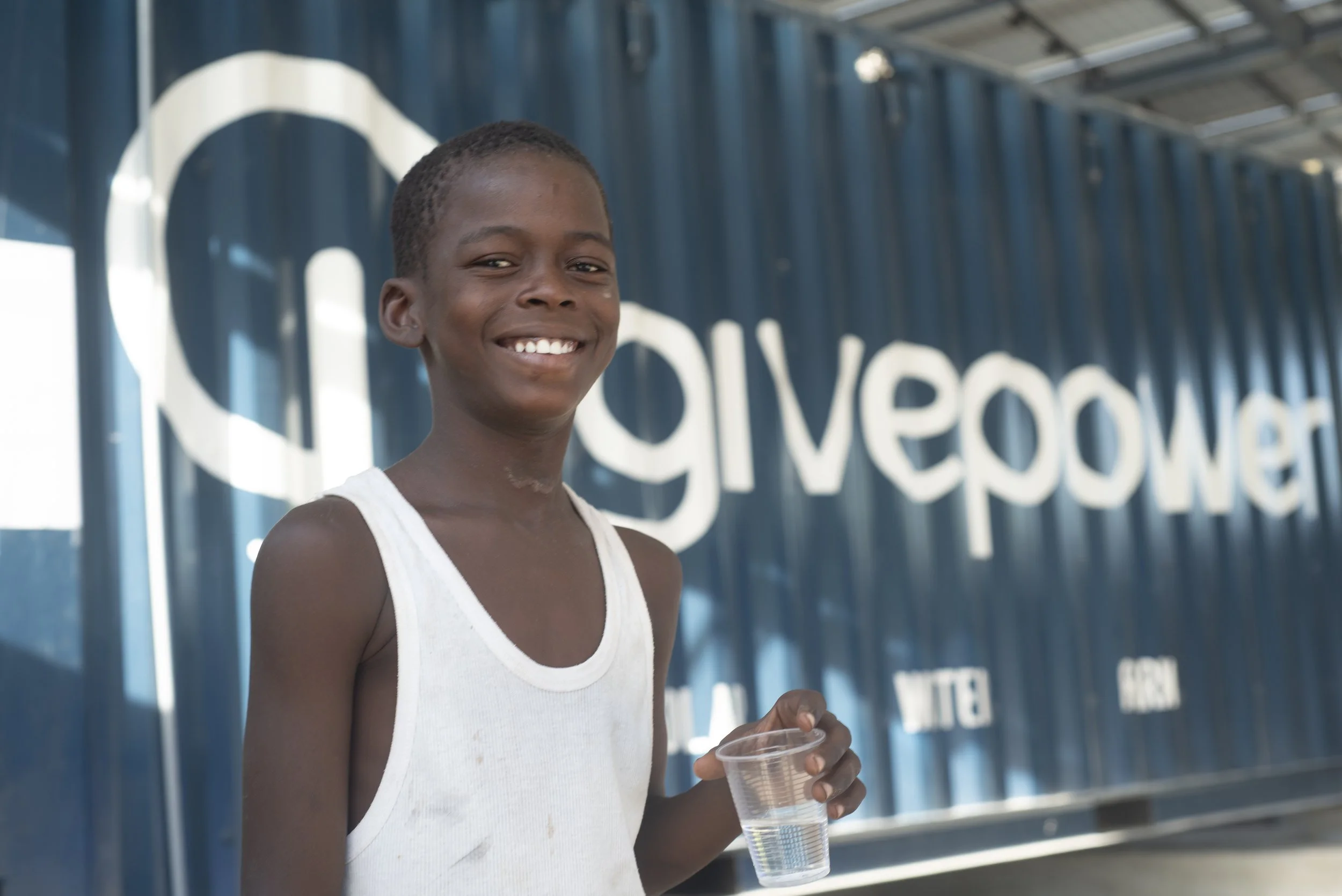 Smiling boy holding a plastic cup of water, standing in front of a blue container with the white text 'givepower' on it.
