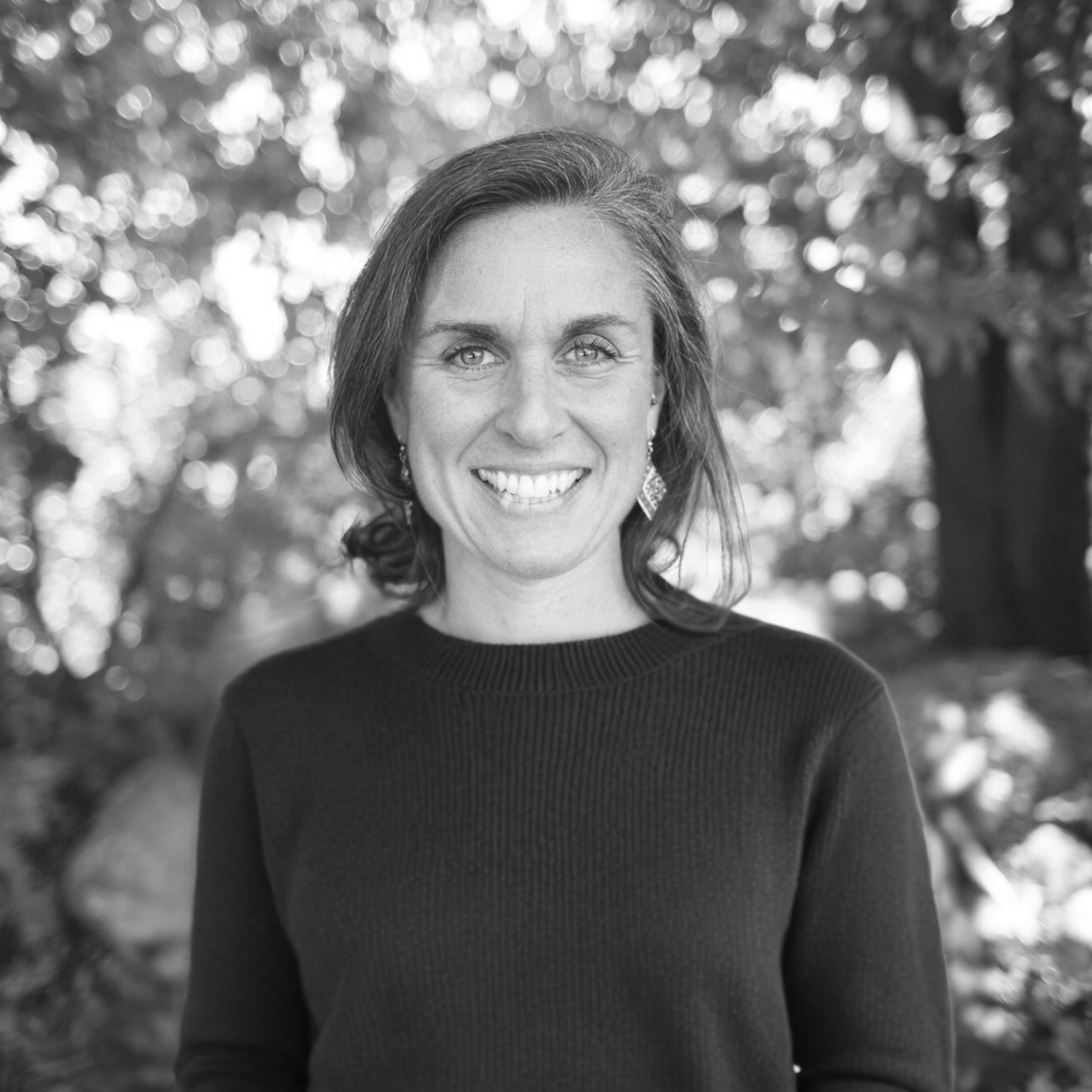Black and white portrait of a smiling woman with shoulder-length hair, wearing earrings and a dark top, outdoors with blurred trees in the background.