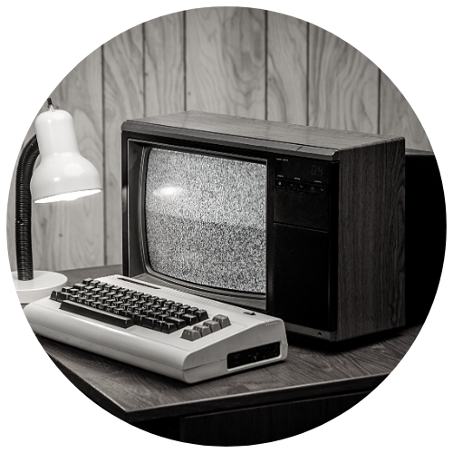 Black and white close up of an old 1990s computer on a desk with a lamp and wood paneling background to symbolize the 1980s computer programming expertise.
