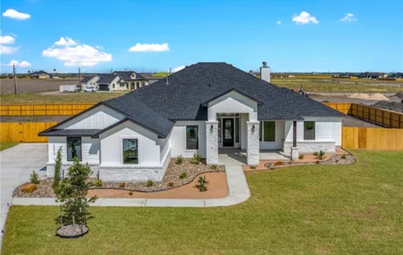 Line drawing of a large house with a gabled roof, multiple windows with shutters, and a central front door. The house features brick or stone accents on the lower exterior and vertical siding on the upper portions.