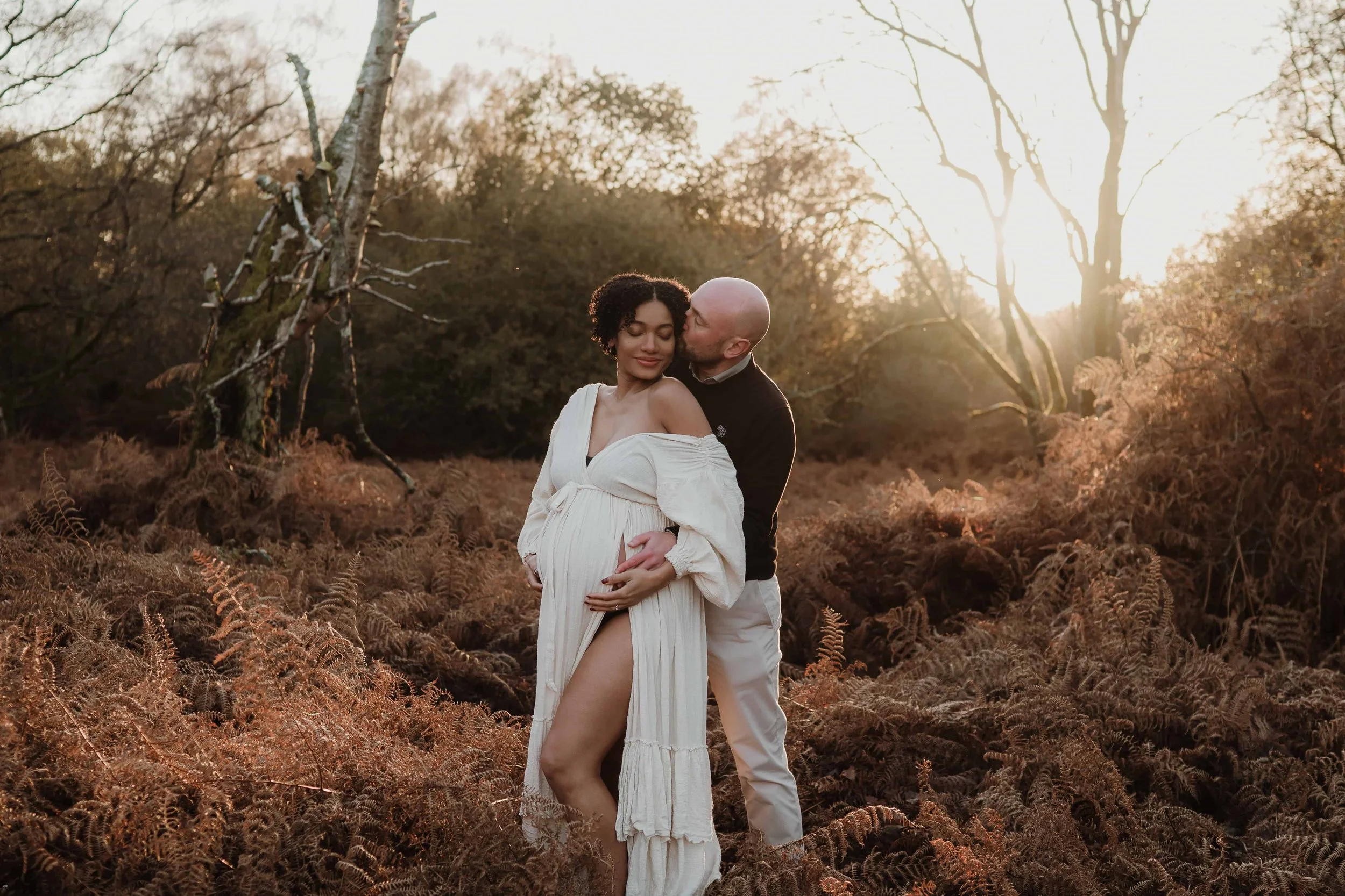 A pregnant woman in a white flowing dress stands in a field of ferns during sunset, being kissed on the cheek by a man in a black sweater and beige pants, with trees and the setting sun in the background.