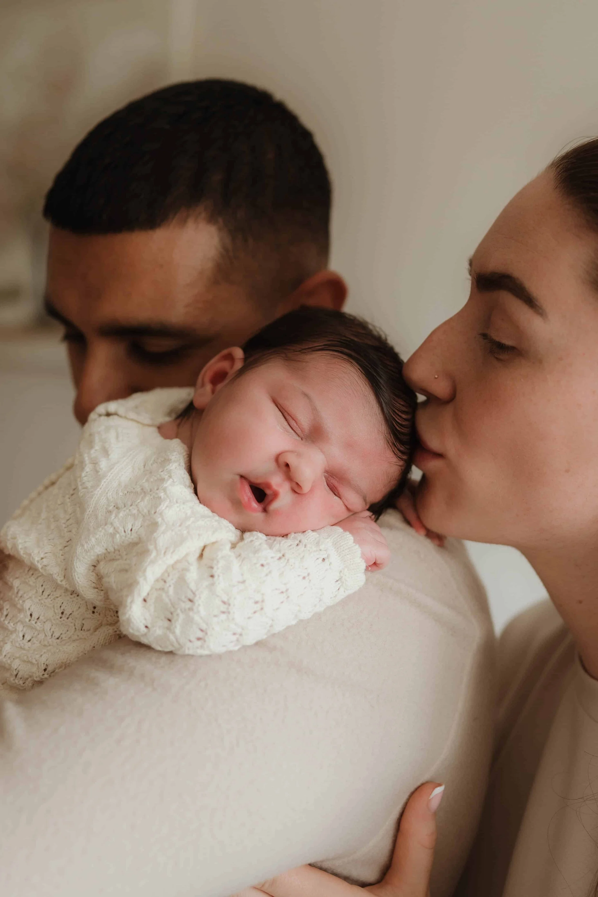 A newborn baby is being held and kissed on the cheek by two adults, one man and one woman, in a loving embrace.