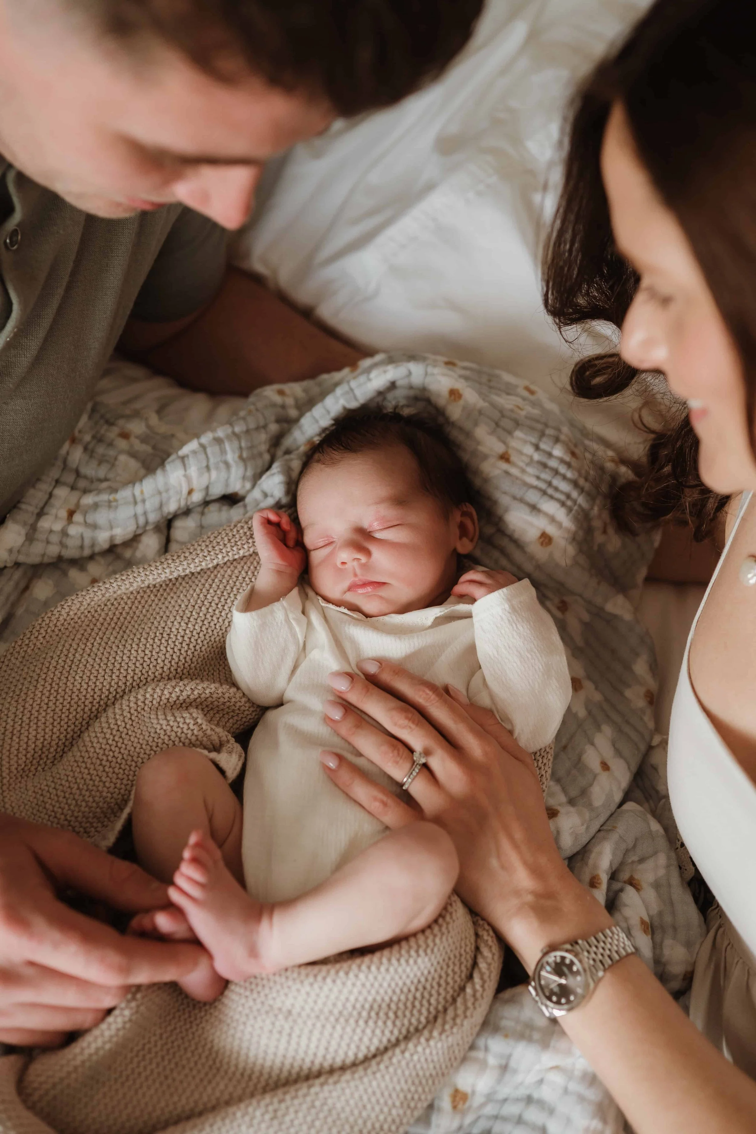 A newborn baby lying in a blanket, surrounded by two adults looking at and holding the baby gently.