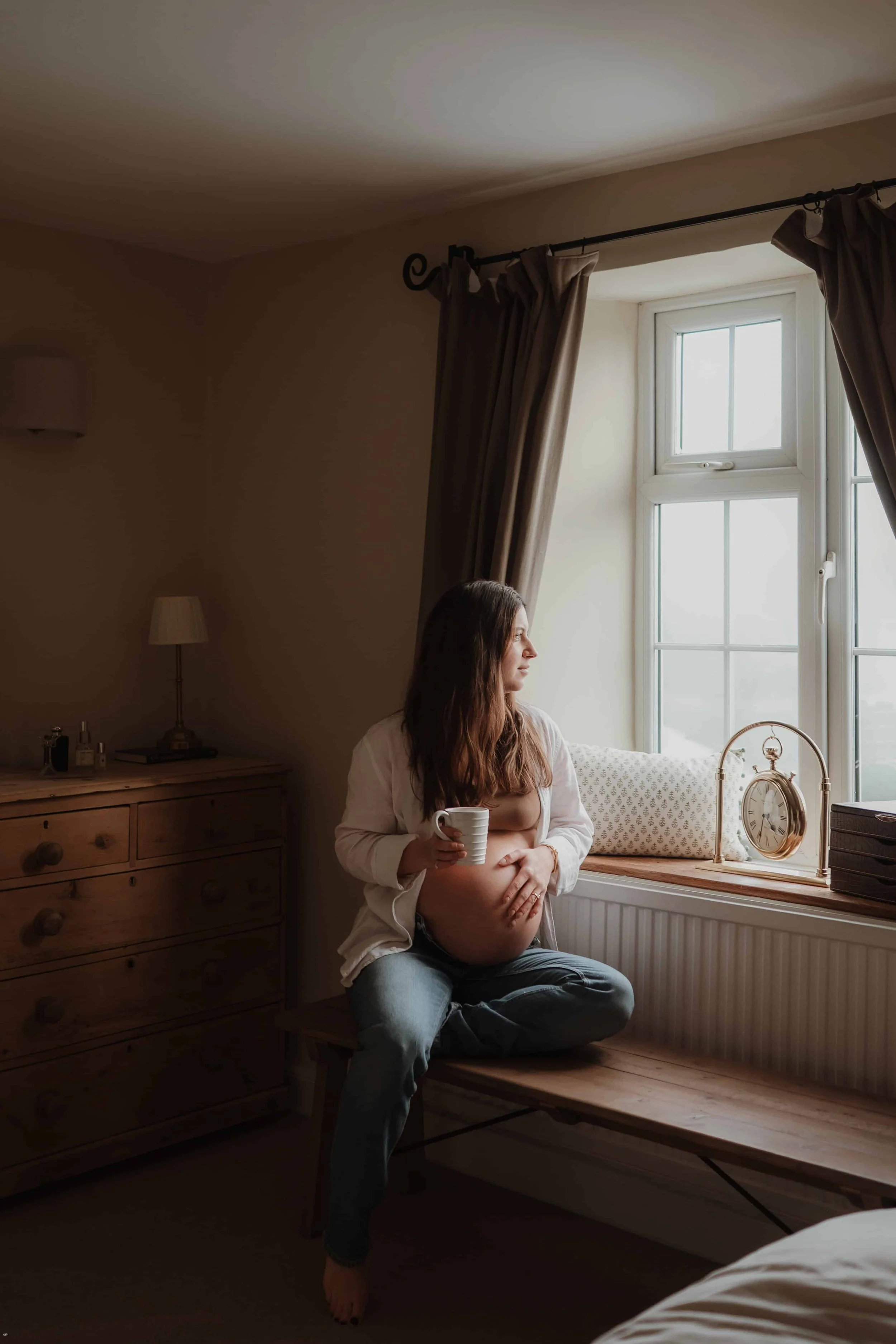 A pregnant woman sitting on a wooden bench by a window, holding a mug and looking outside, in a softly lit bedroom with beige walls, chasing curtains, a wooden dresser with a lamp, and a clock on the windowsill.