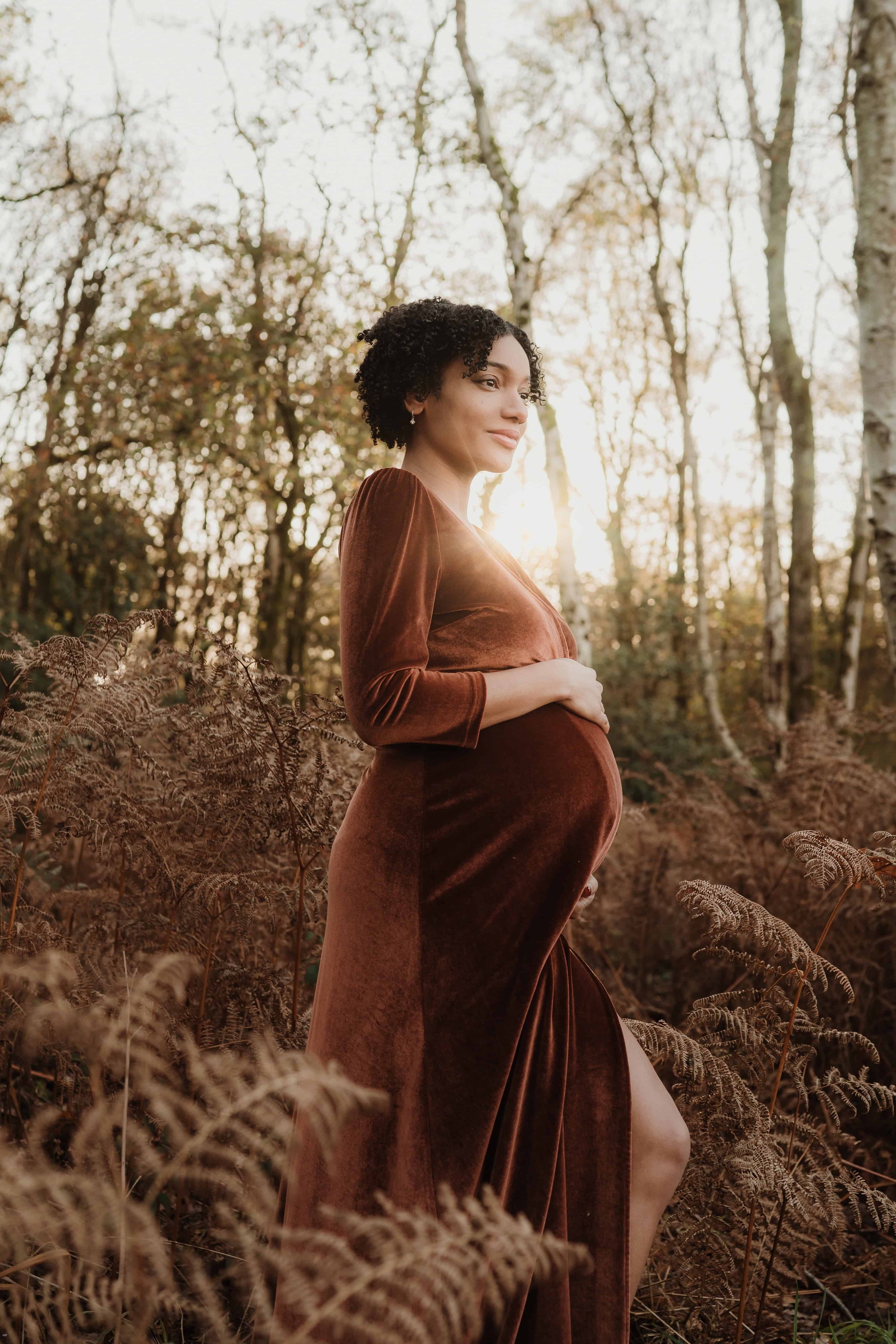 A pregnant woman in a brown velvet dress standing in a forest with trees and ferns, sunlight shining behind her.