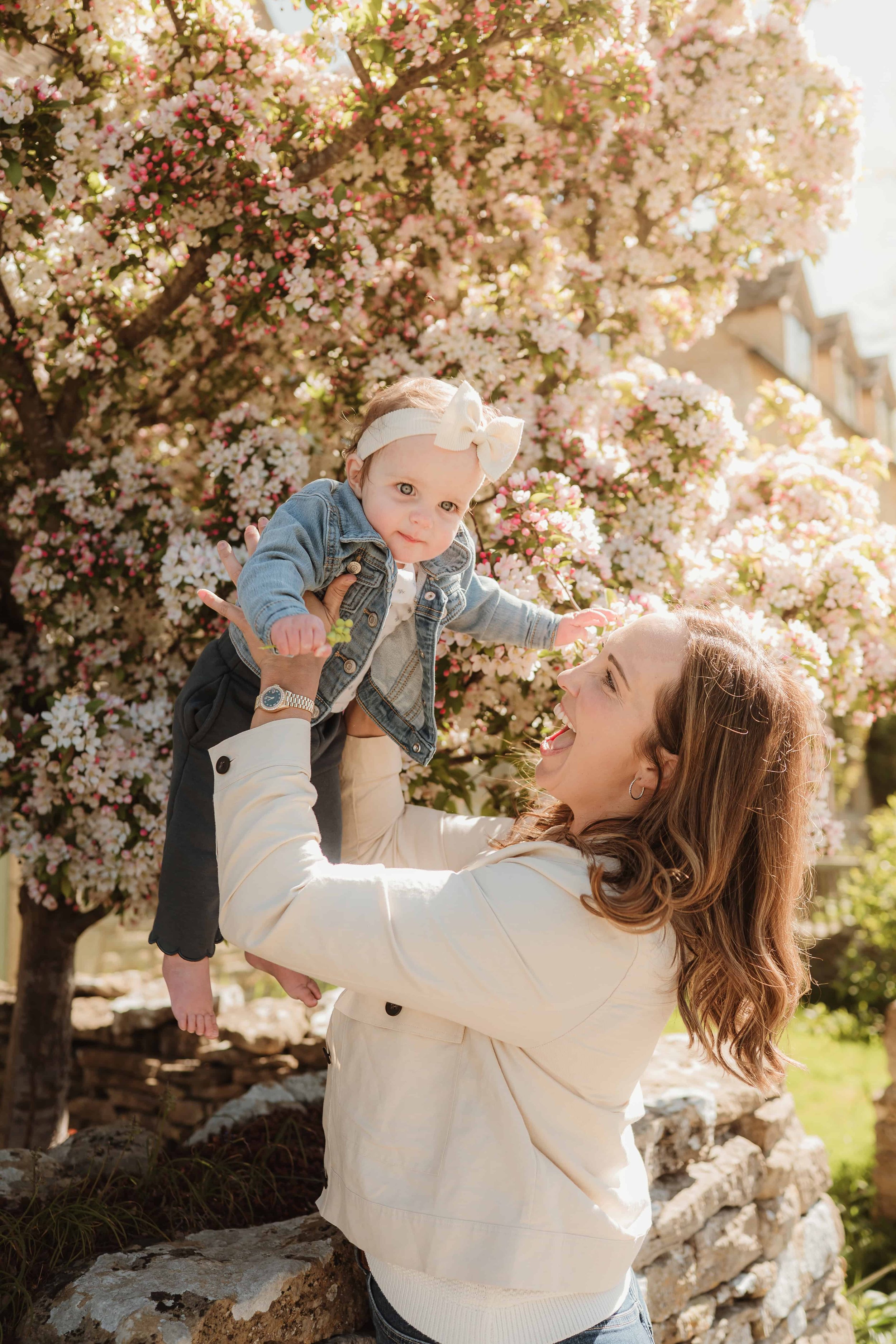 A woman with long wavy brown hair smiling and looking up as she lifts a baby girl in front of a flowering tree with pink and white blossoms.