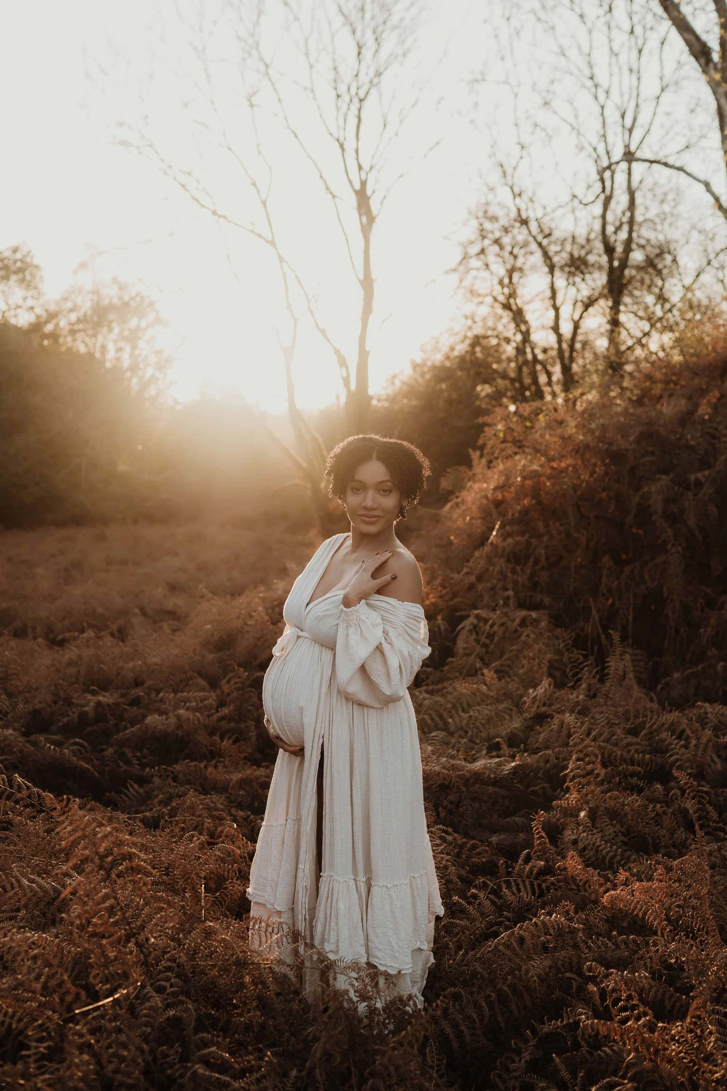 A pregnant woman with curly hair stands in a field of ferns during sunset. She wears a flowing beige dress, cradles her belly with one hand, and touches her shoulder with the other. The sun is setting behind her, creating a warm glow and highlighting
