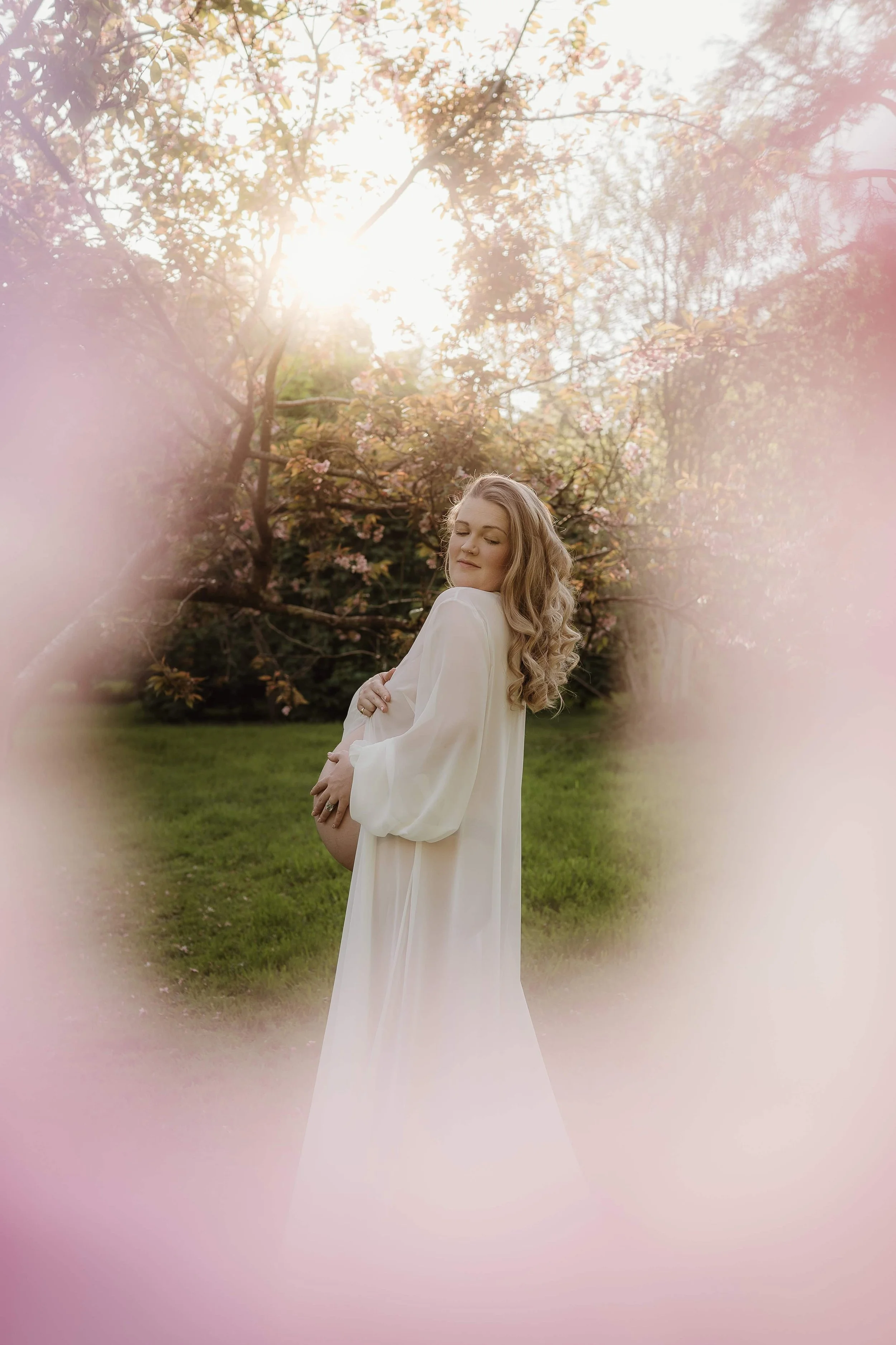 A pregnant woman with long, curly blonde hair standing outdoors by blooming pink cherry blossom trees, with the sun shining through the branches, wearing a flowing white dress.