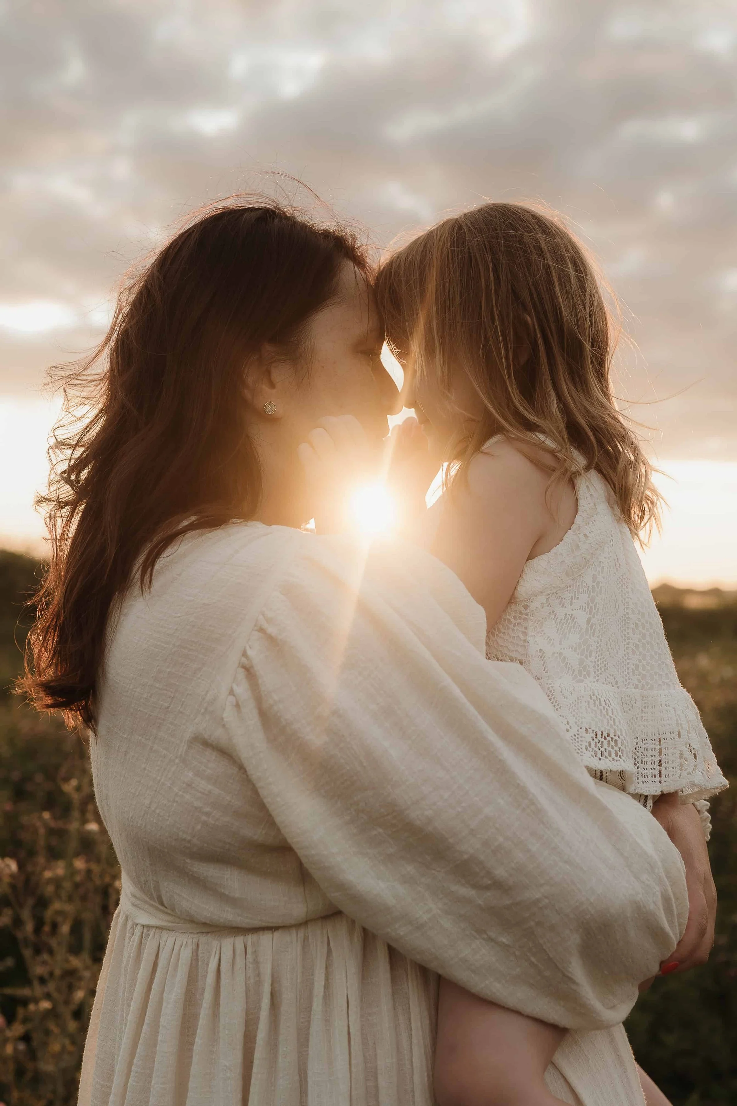 A mother holding her young daughter close outdoors during sunset, with their foreheads touching and sunlight shining between them.