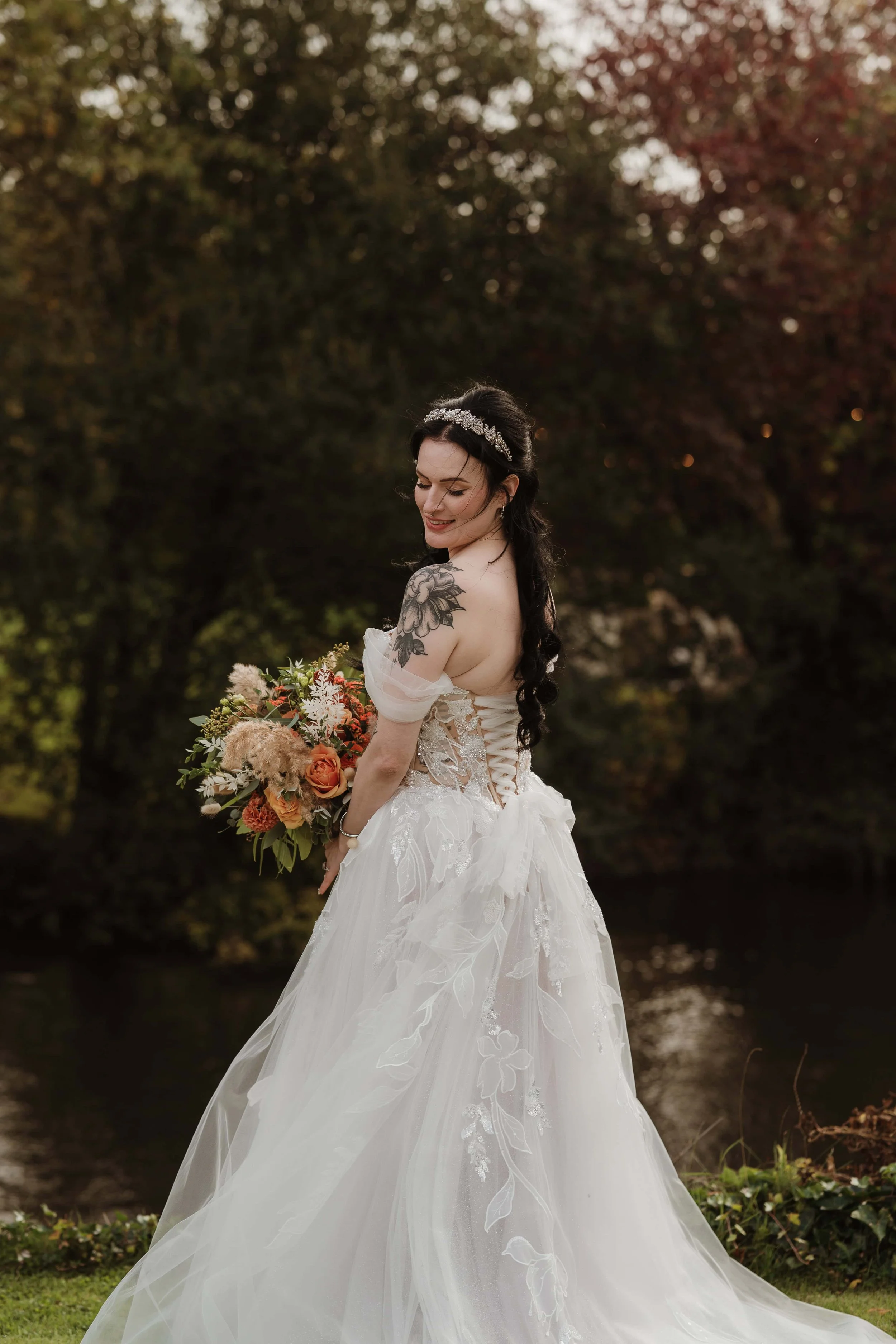 A bride in a white wedding dress holding a bouquet of flowers outdoors near a river, with trees in the background during sunset.