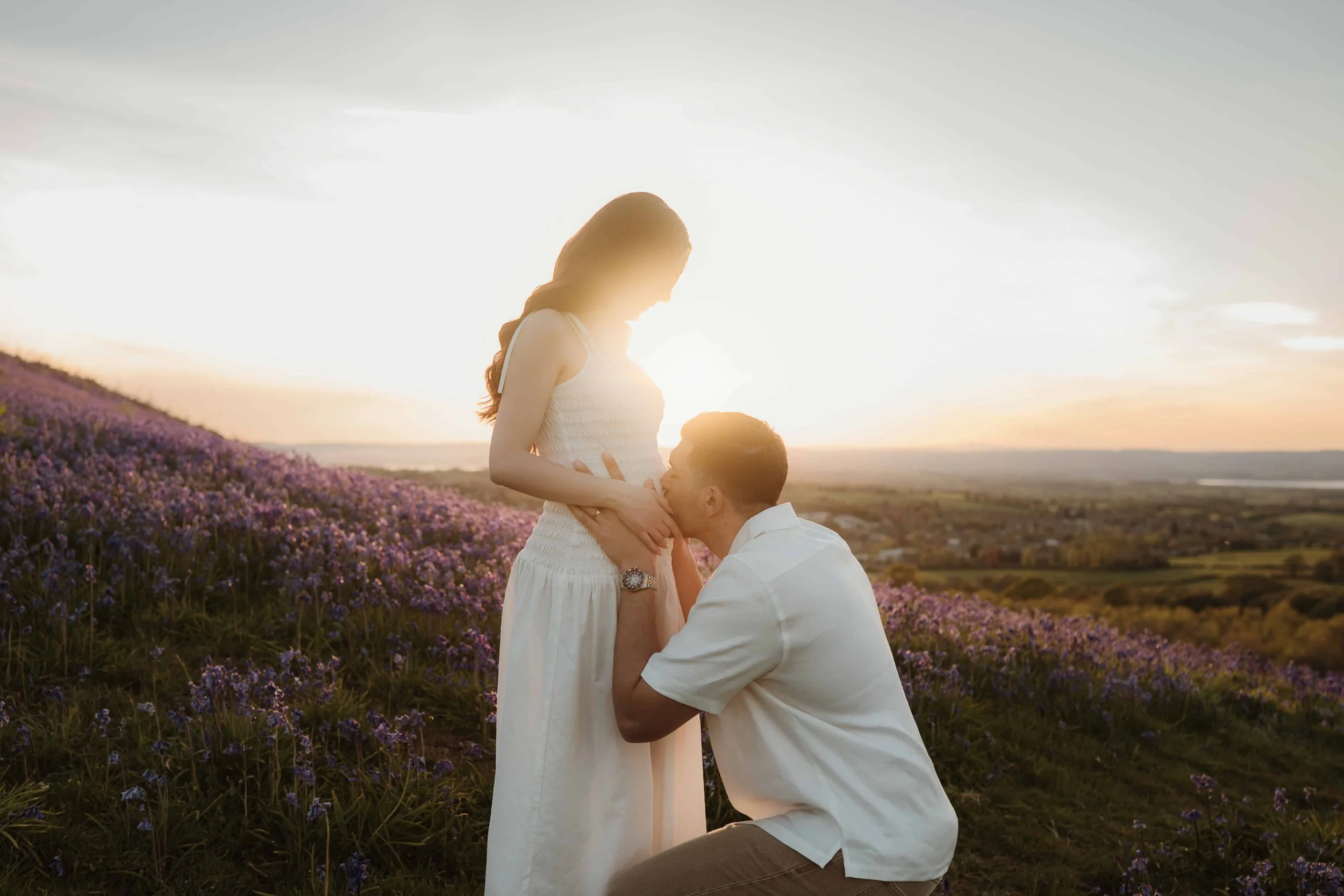 A man kneeling and kissing a pregnant woman on her belly in a field of purple flowers during sunset.