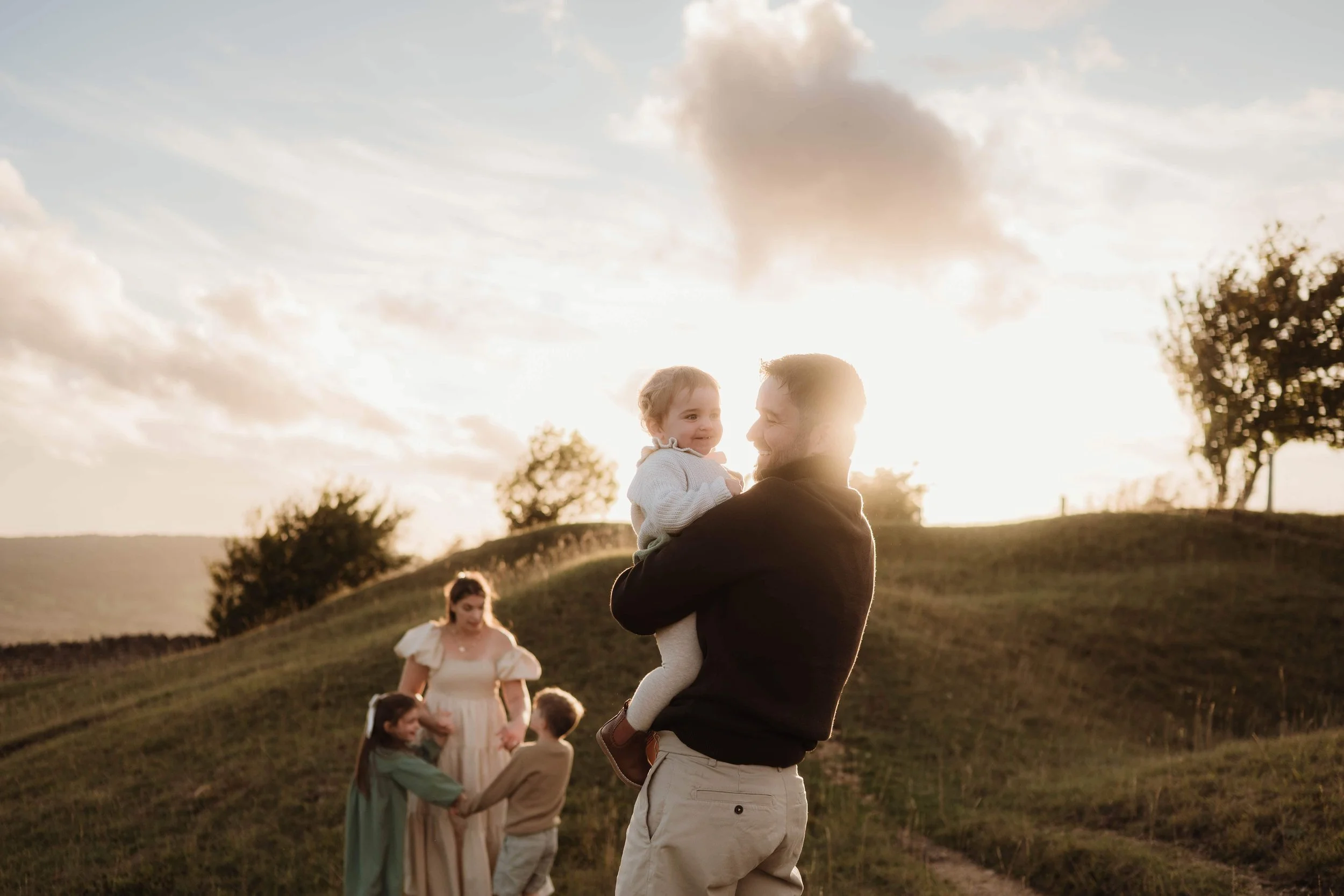 a family of five outdoors at sunset on crickley hill in cheltenham. Dad is holding the toddler while mum dances with the other two children
