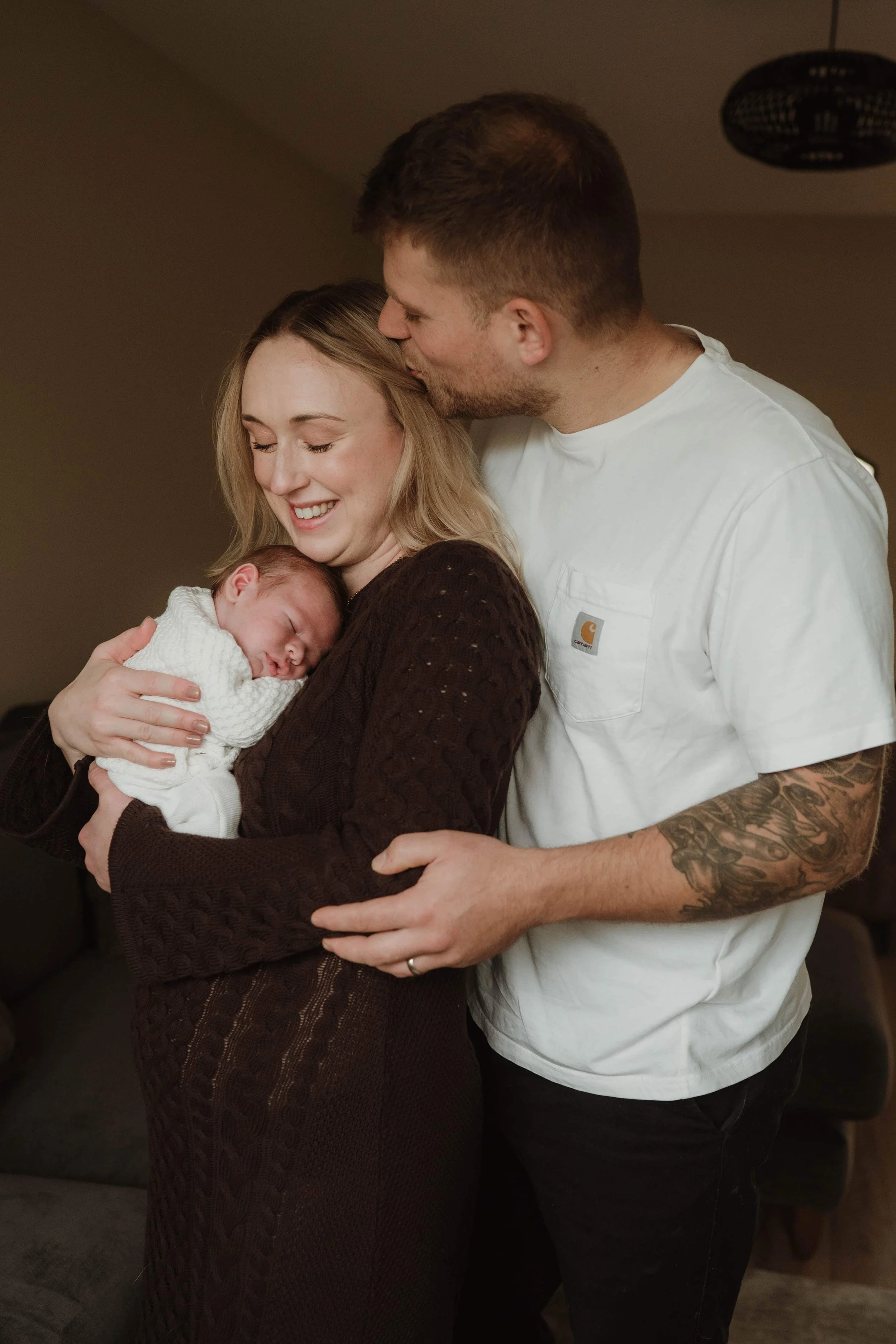 A couple holding a newborn baby indoors, with the man kissing the woman's forehead. The woman is smiling with her eyes closed, and the baby is wrapped in a white blanket, sleeping on her chest.