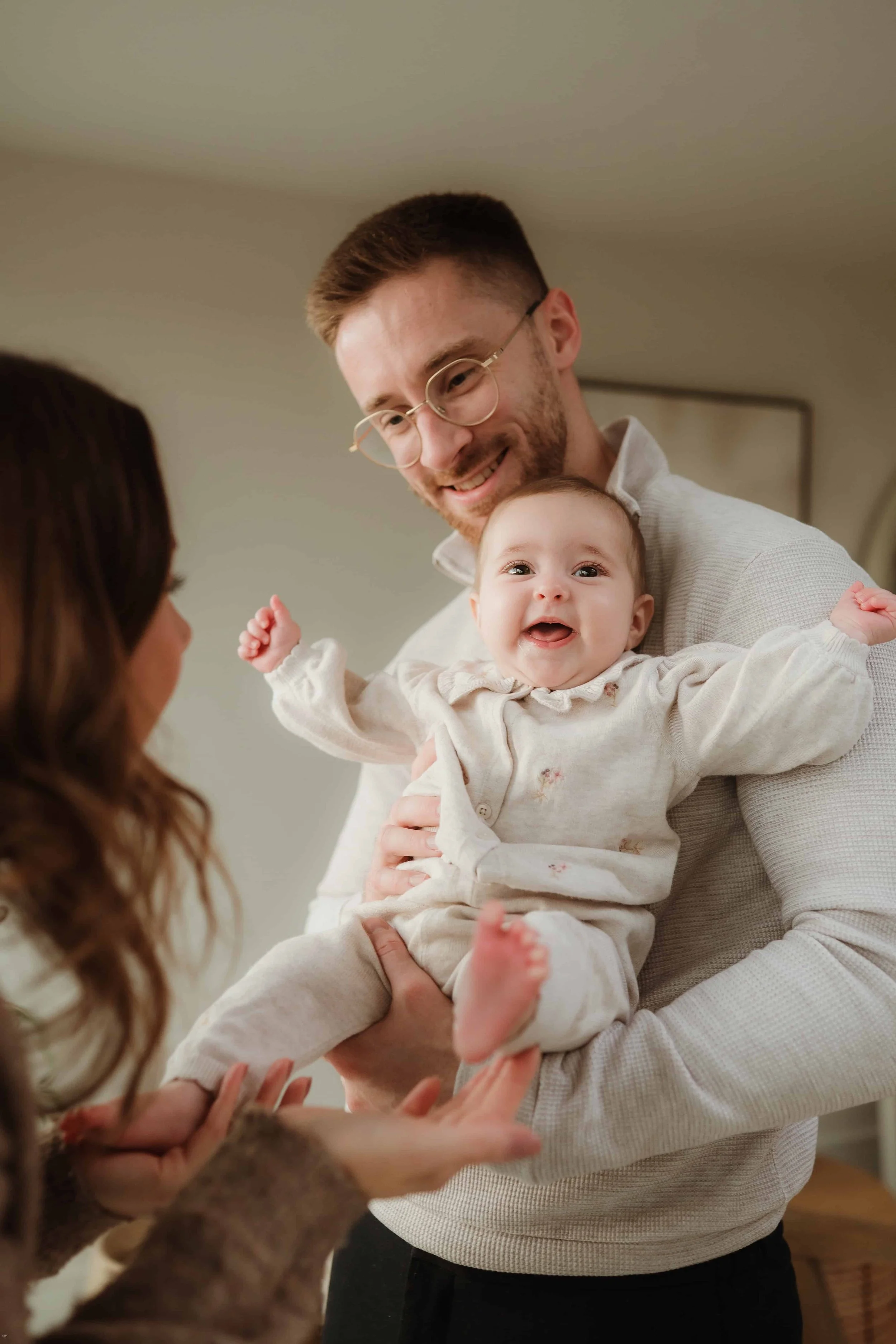 A man with glasses and a beard holds a happy baby with a big smile, while a woman with long curly hair reaches out toward the baby.