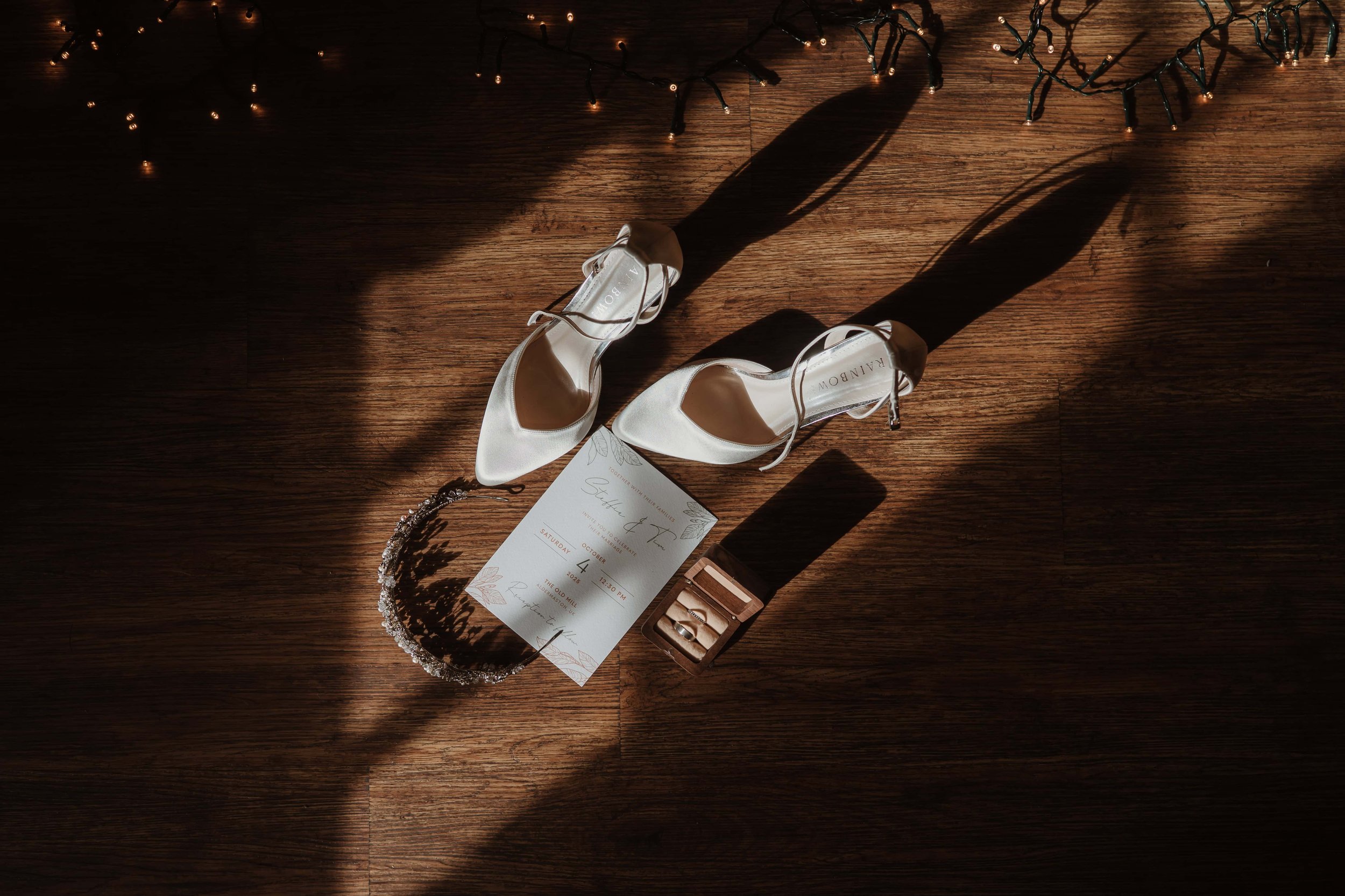 A pair of white satin high-heeled shoes, a wedding invitation, a tiara, and a small ring box with rings on a wooden floor, with shadows cast by the sunlight.