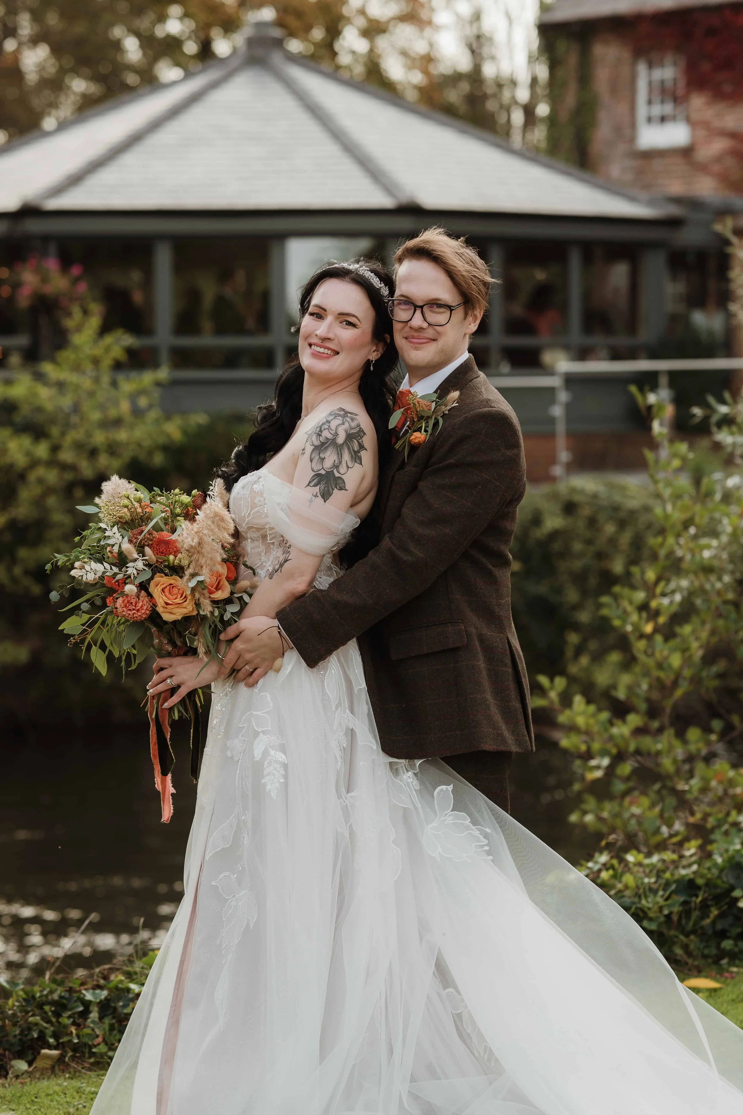 A newlywed couple standing outdoors near a pond, smiling and embracing. The bride is wearing a white wedding dress with off-the-shoulder sleeves and holding a bouquet of flowers. The groom is dressed in a brown jacket and glasses, with a boutonniere. The background features a garden with trees, shrubs, and a building with a porch.