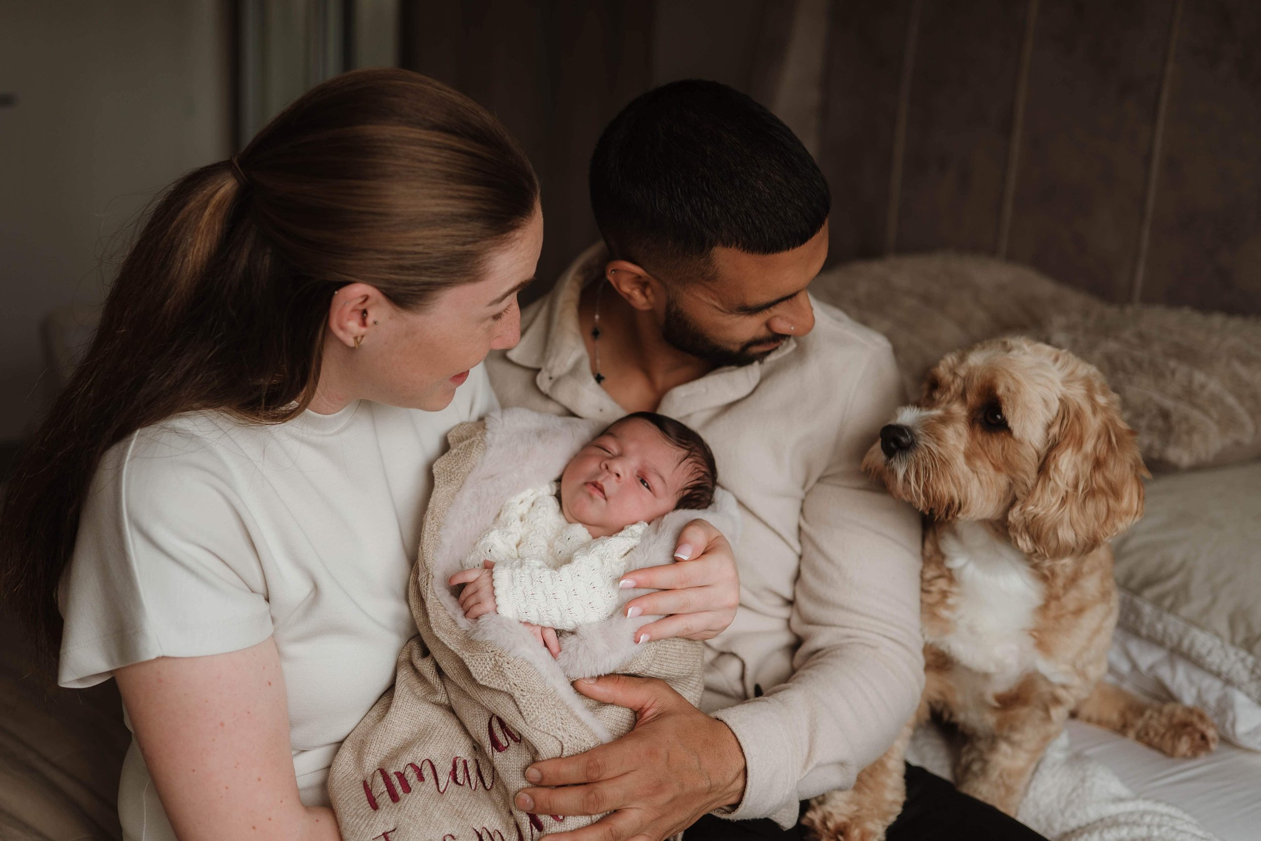 Family of three, a man, woman, and their newborn baby, with a dog, sitting on a bed in a cozy room.