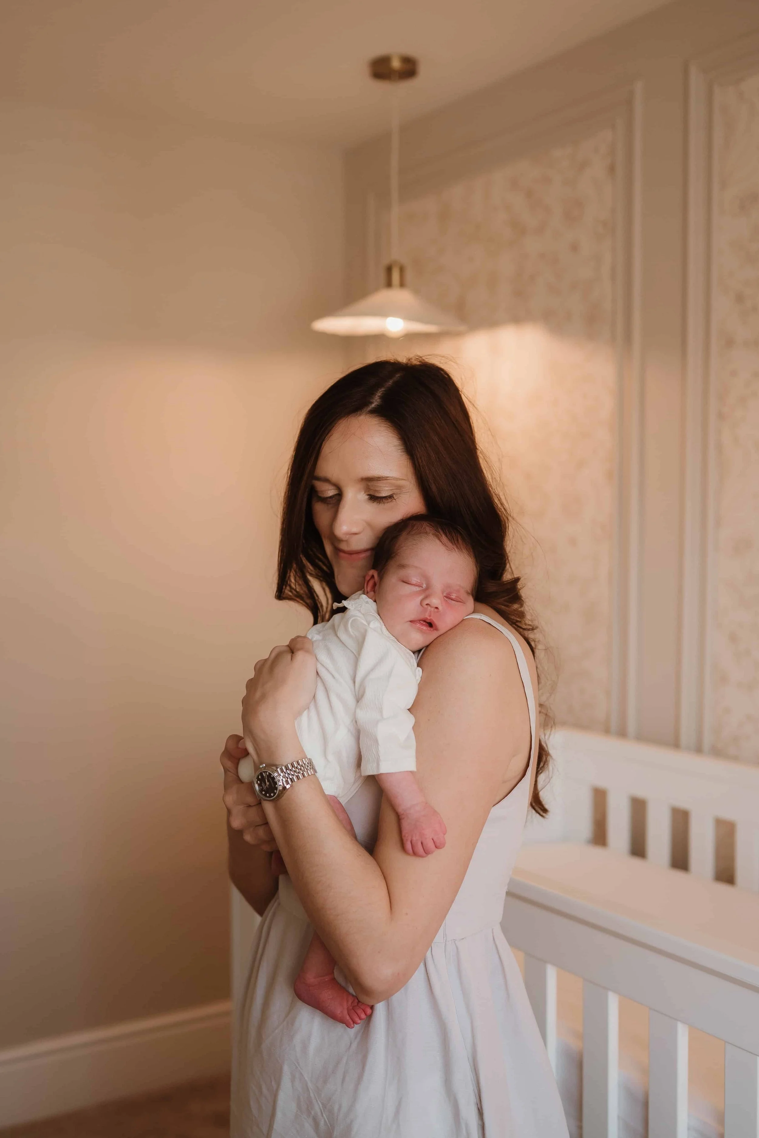 A woman with dark brown hair holding a sleeping baby in a light-colored dress inside a room with soft lighting and a beige wall with decorative paneling.