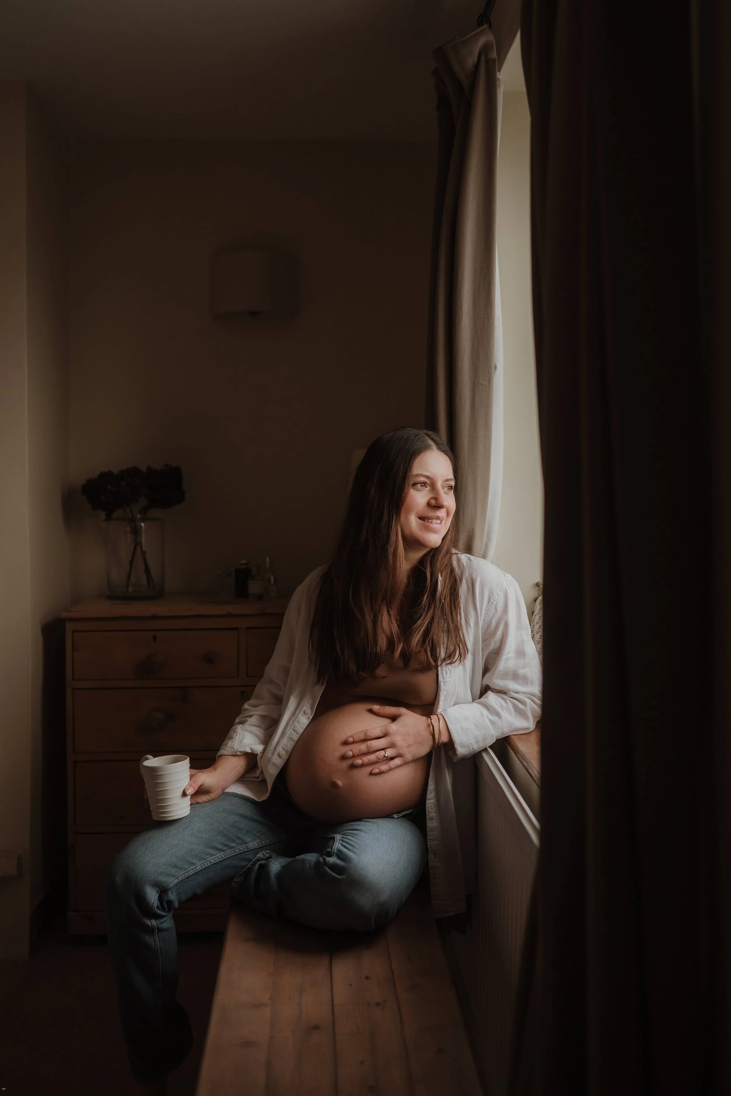 A pregnant woman sitting on a windowsill, holding a mug in one hand, and gently touching her belly with the other, smiling and looking out the window.