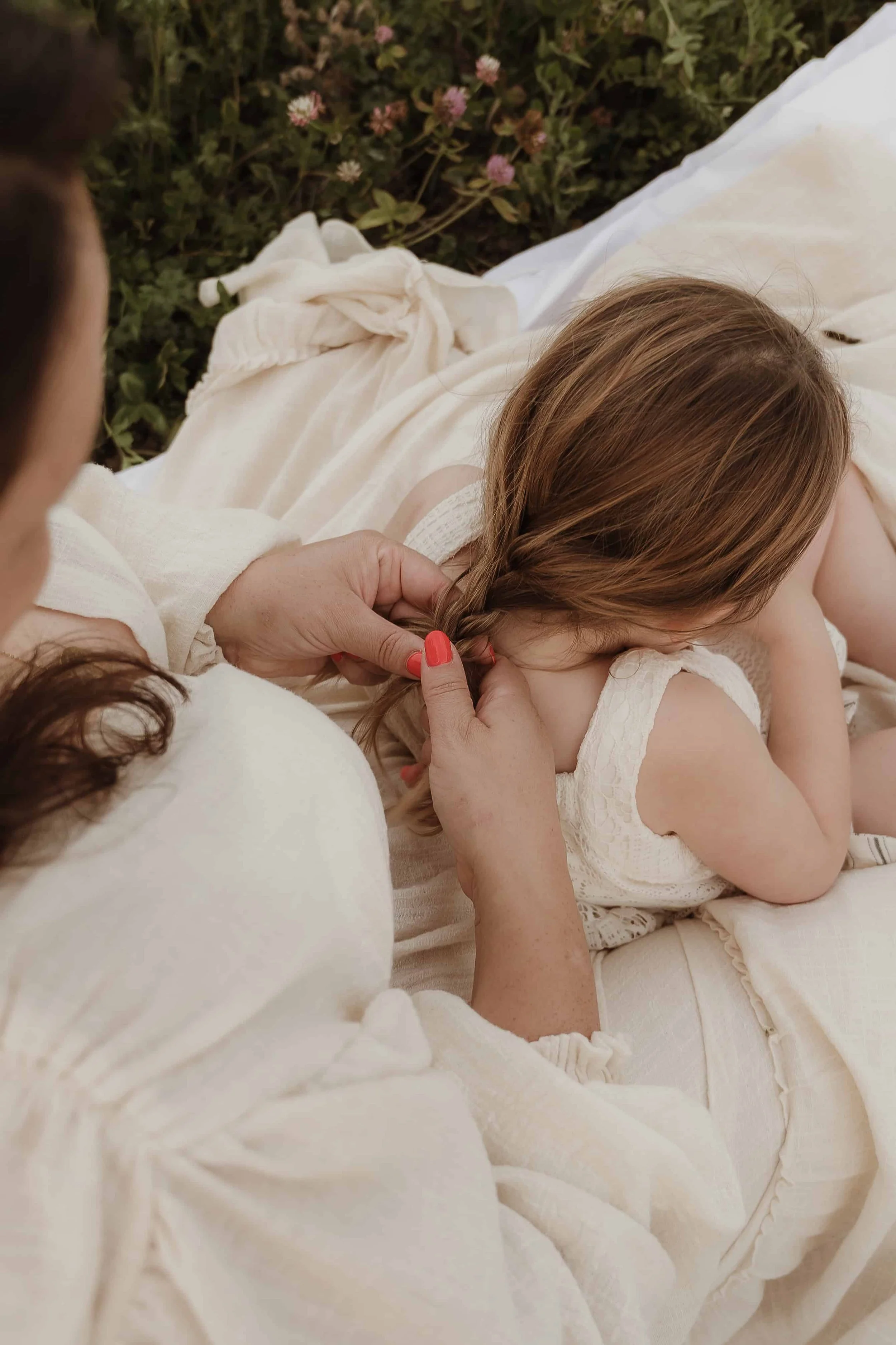 A woman with red nail polish gently holding and brushing a young girl's long, light brown hair outside on a blanket near flowers.