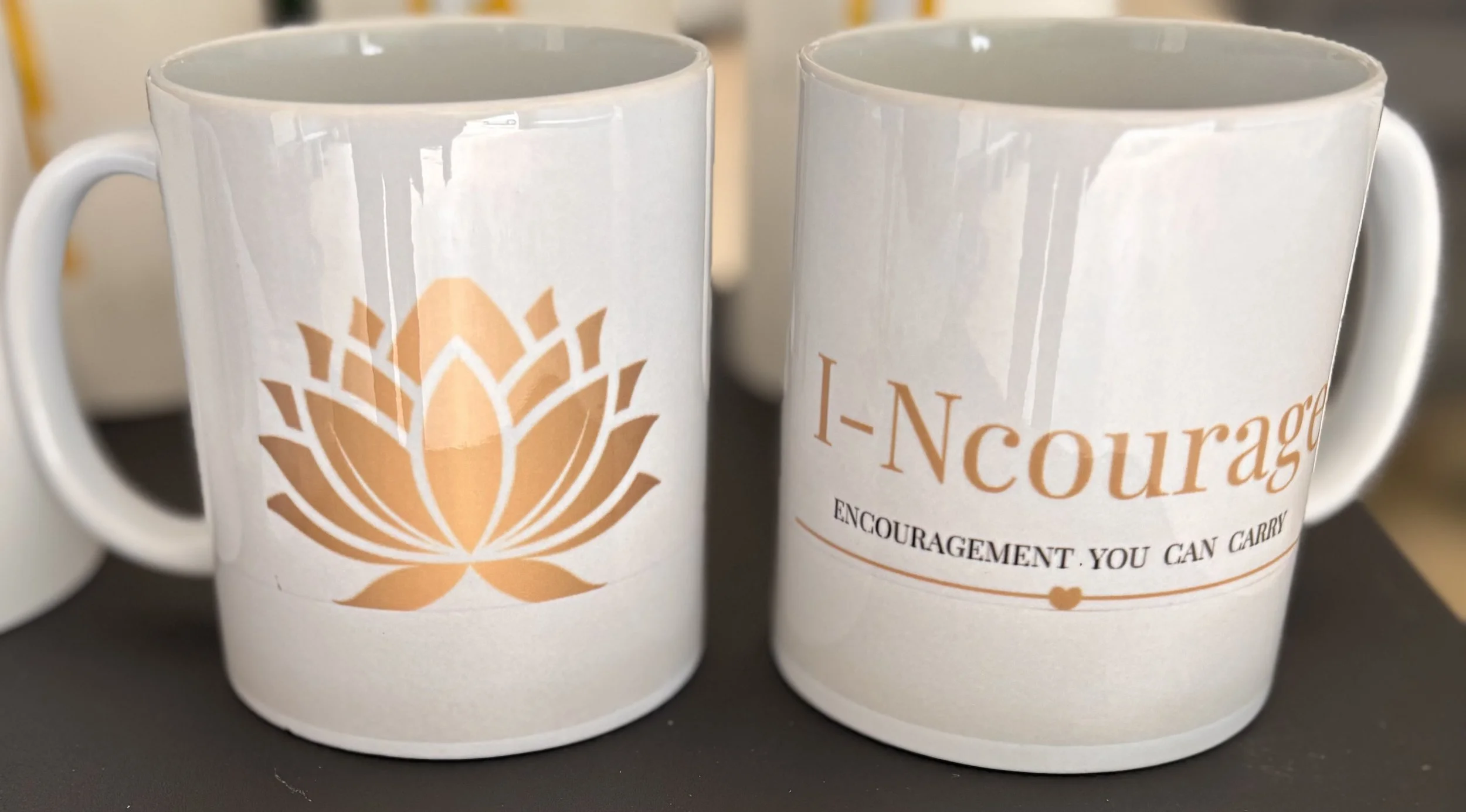 Two white ceramic mugs with printed text and a logo in gold. One mug has a lotus-like design, and the other has the word 'I-Ncourage' and additional text that says Encouragement You Can Carry.