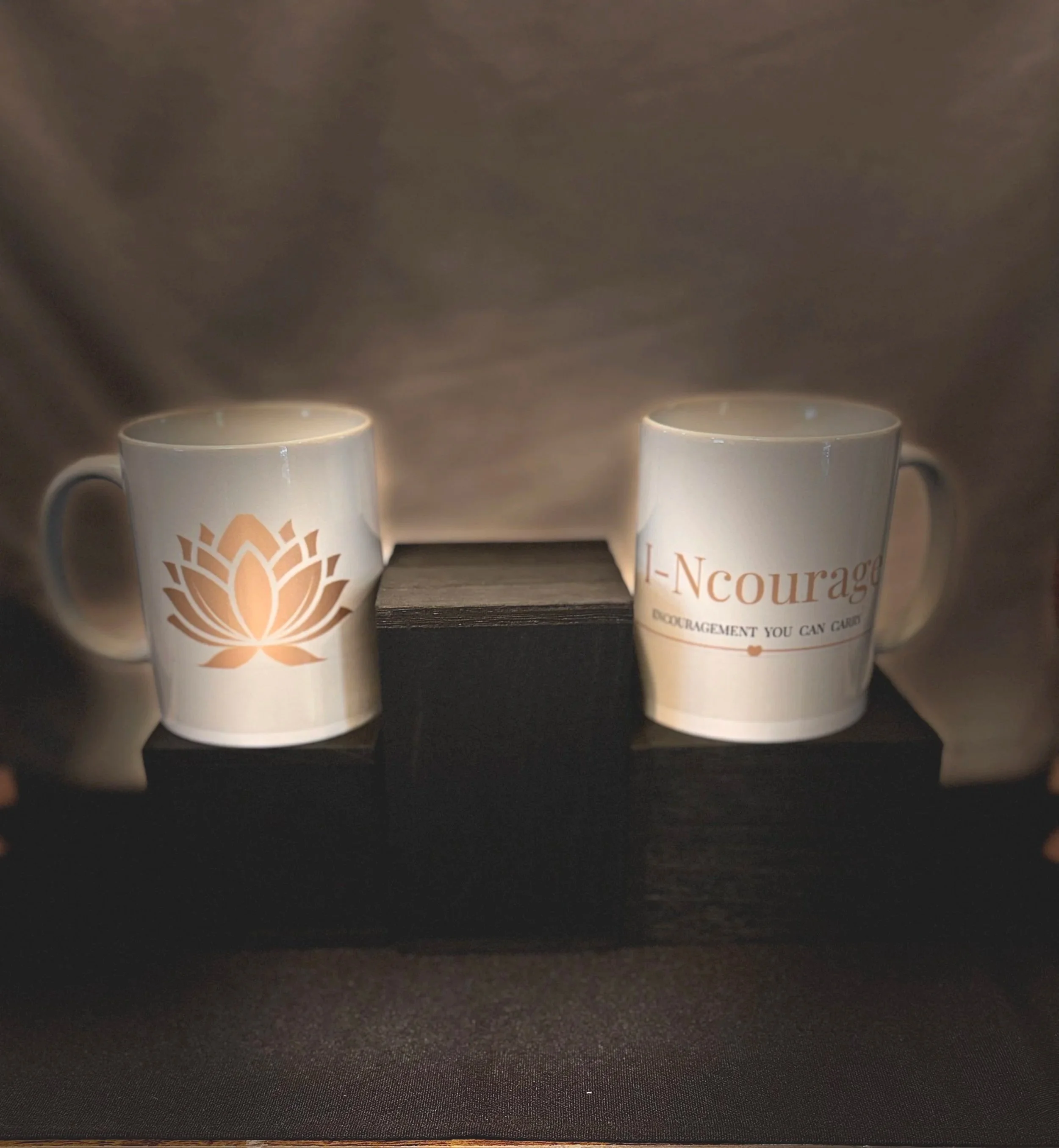 Two white ceramic mugs with a gold logo and text, placed on a black surface against a dark background.