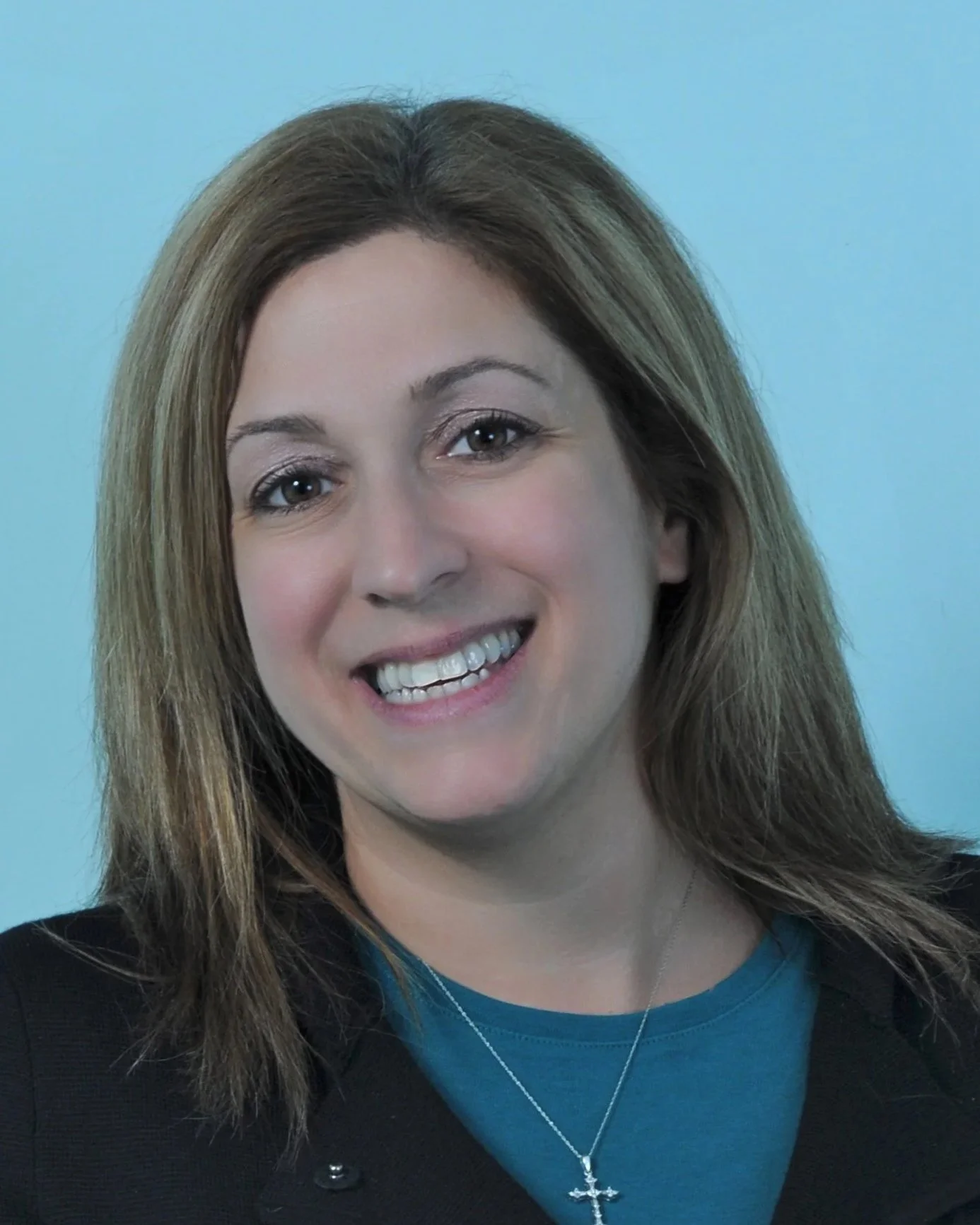 Smiling woman with shoulder-length brown hair, wearing a blue top, a black jacket, and a cross necklace, against a light blue background.