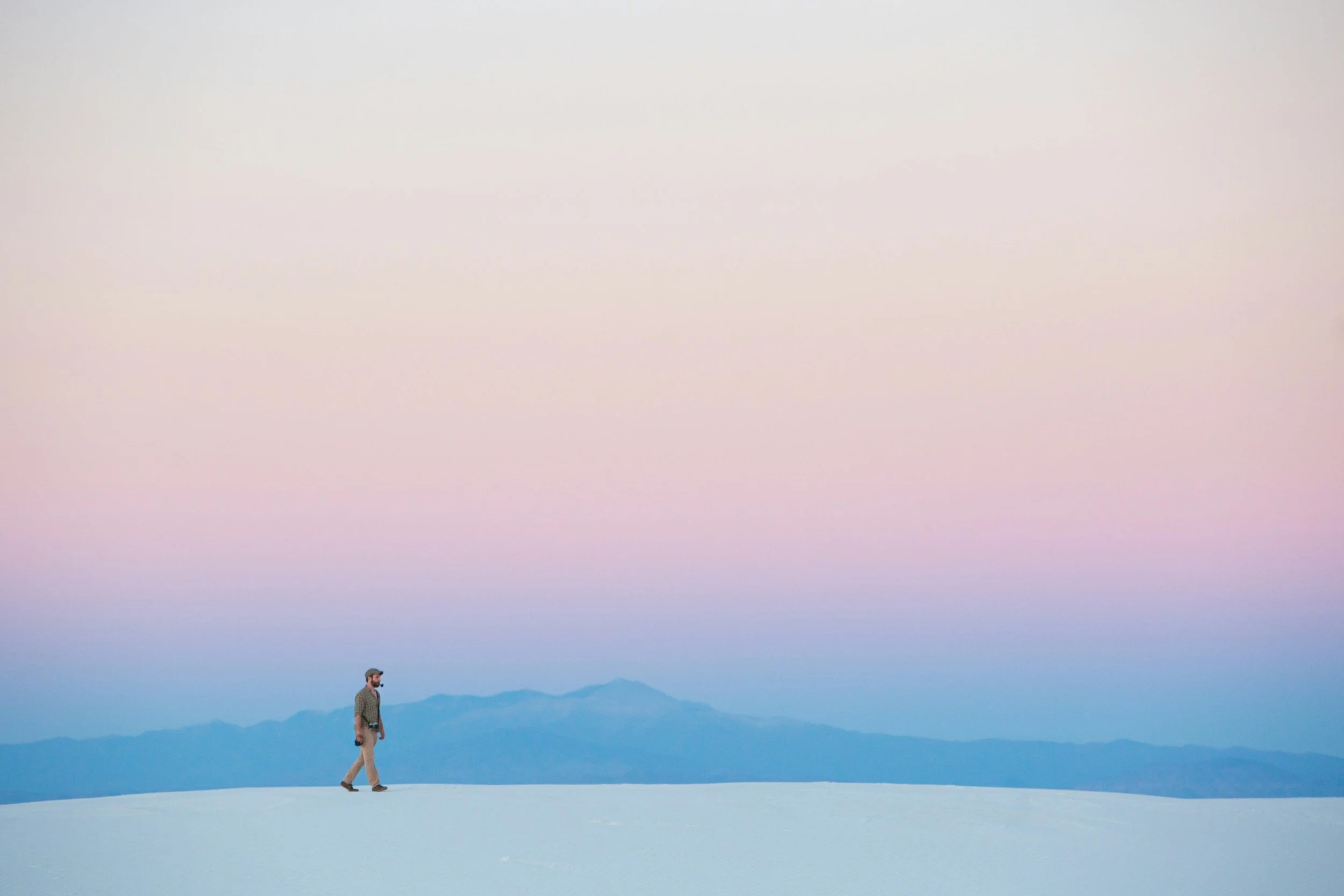 a figure walks across a sandy landscape with pastel mountains and sky behind them