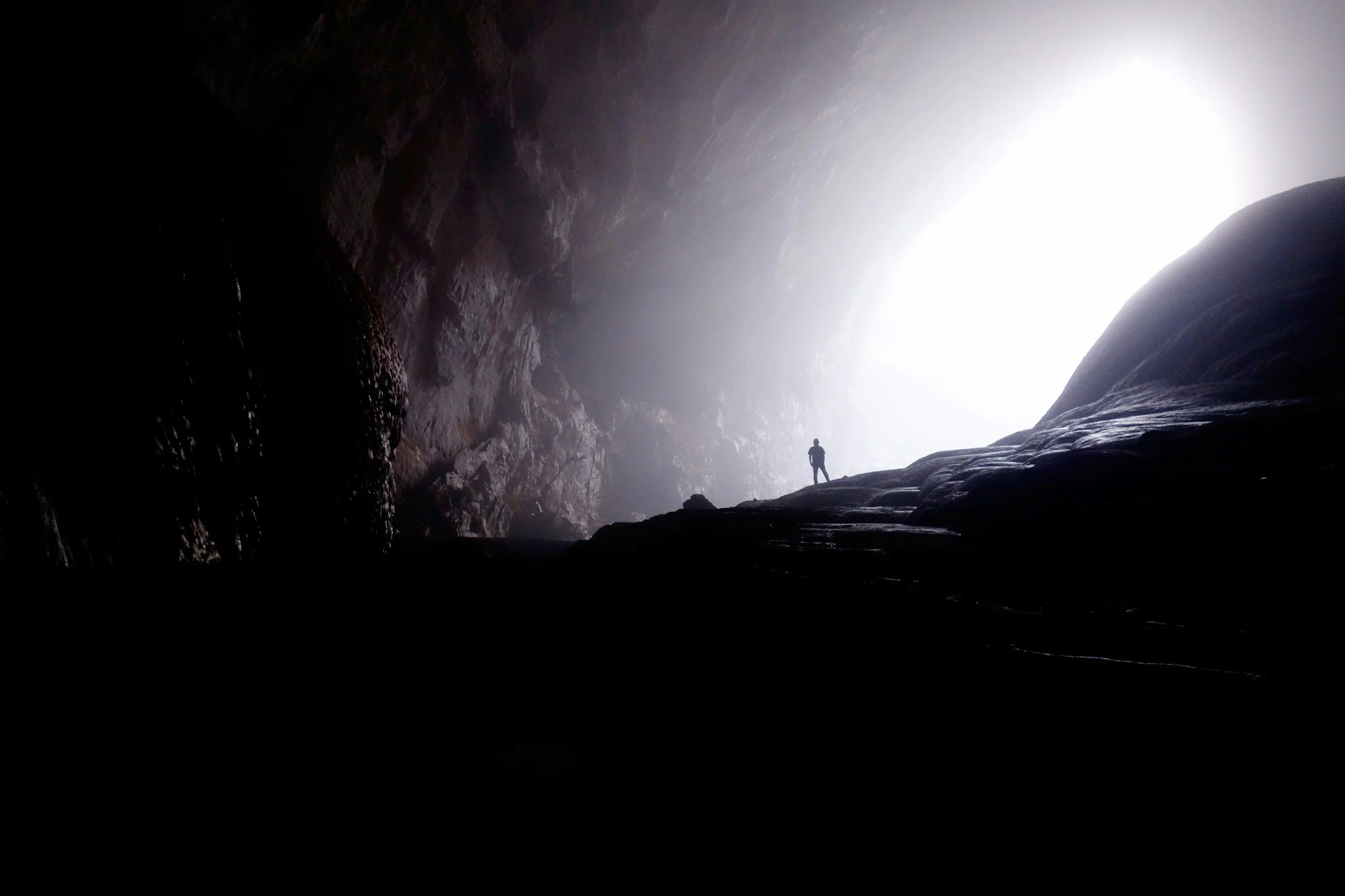 A person stands at the entrance of a cave, light pouring through the cave's opening.
