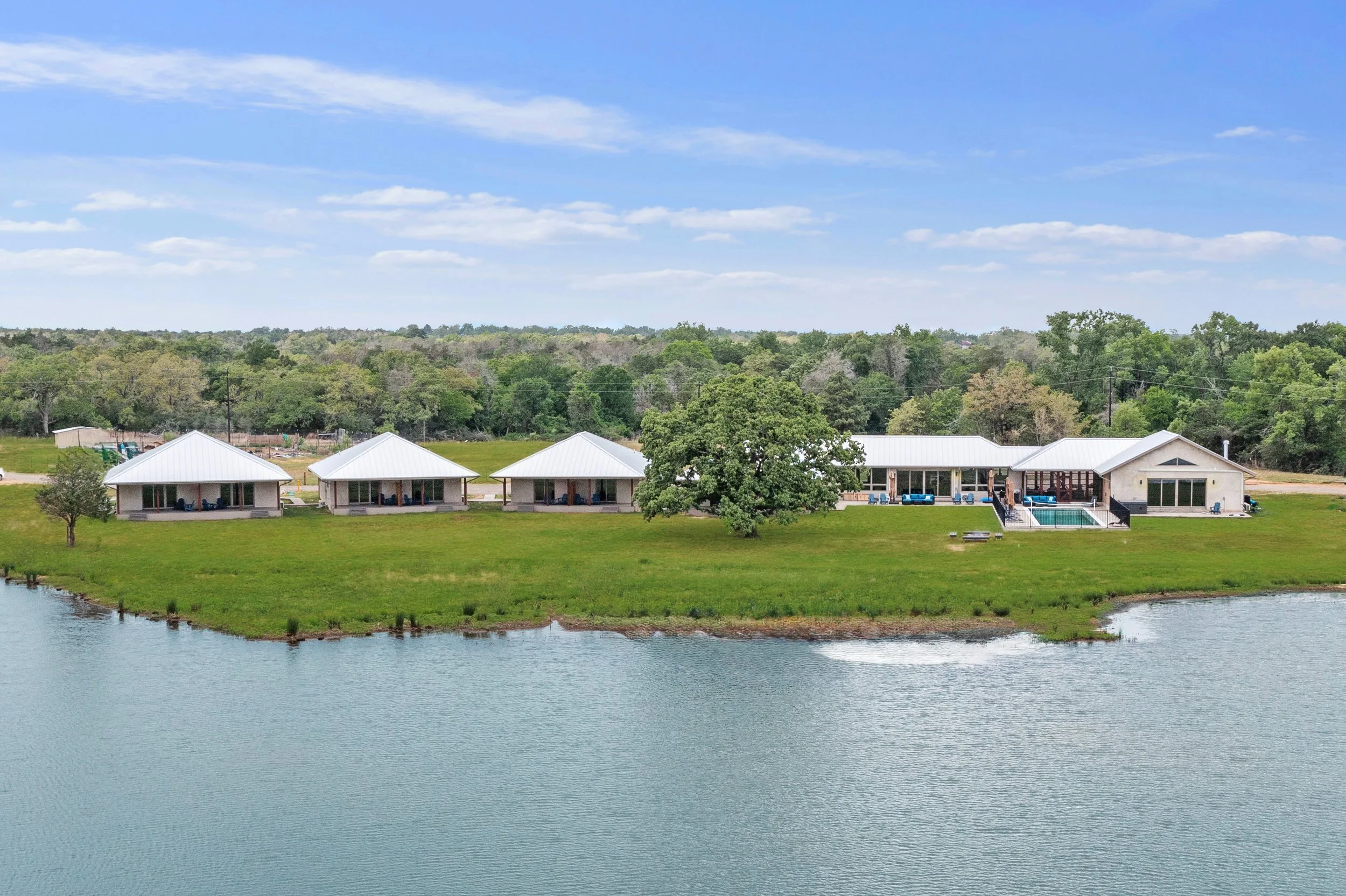 A peaceful lakeside scene with a house and a pool on the shore, surrounded by green trees and grassy land, under a blue sky with scattered clouds.