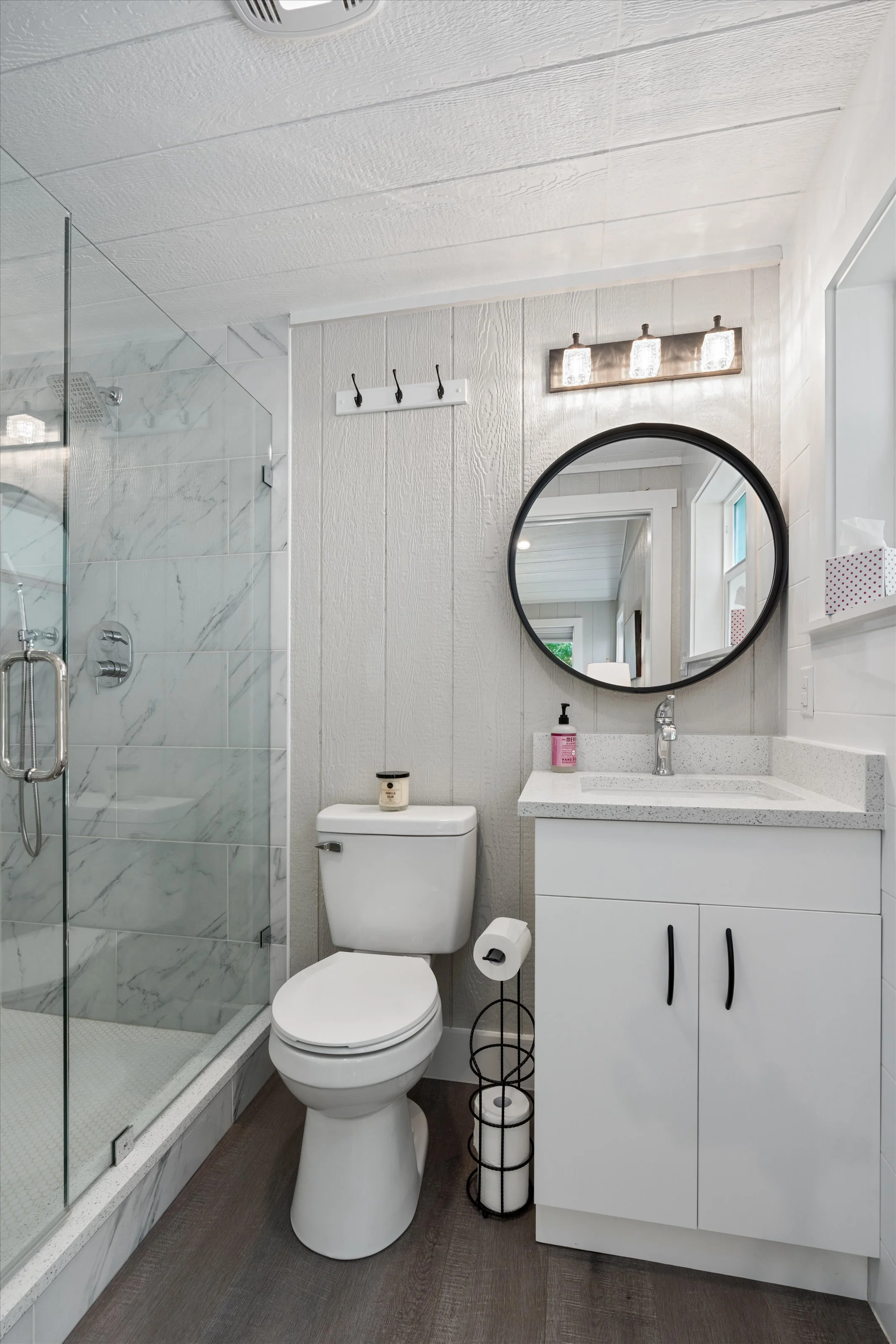 Modern bathroom with walk-in shower, round mirror above sink, white vanity, toilet, and wall-mounted light fixture. Decorative wire basket and small items on the vanity and toilet.
