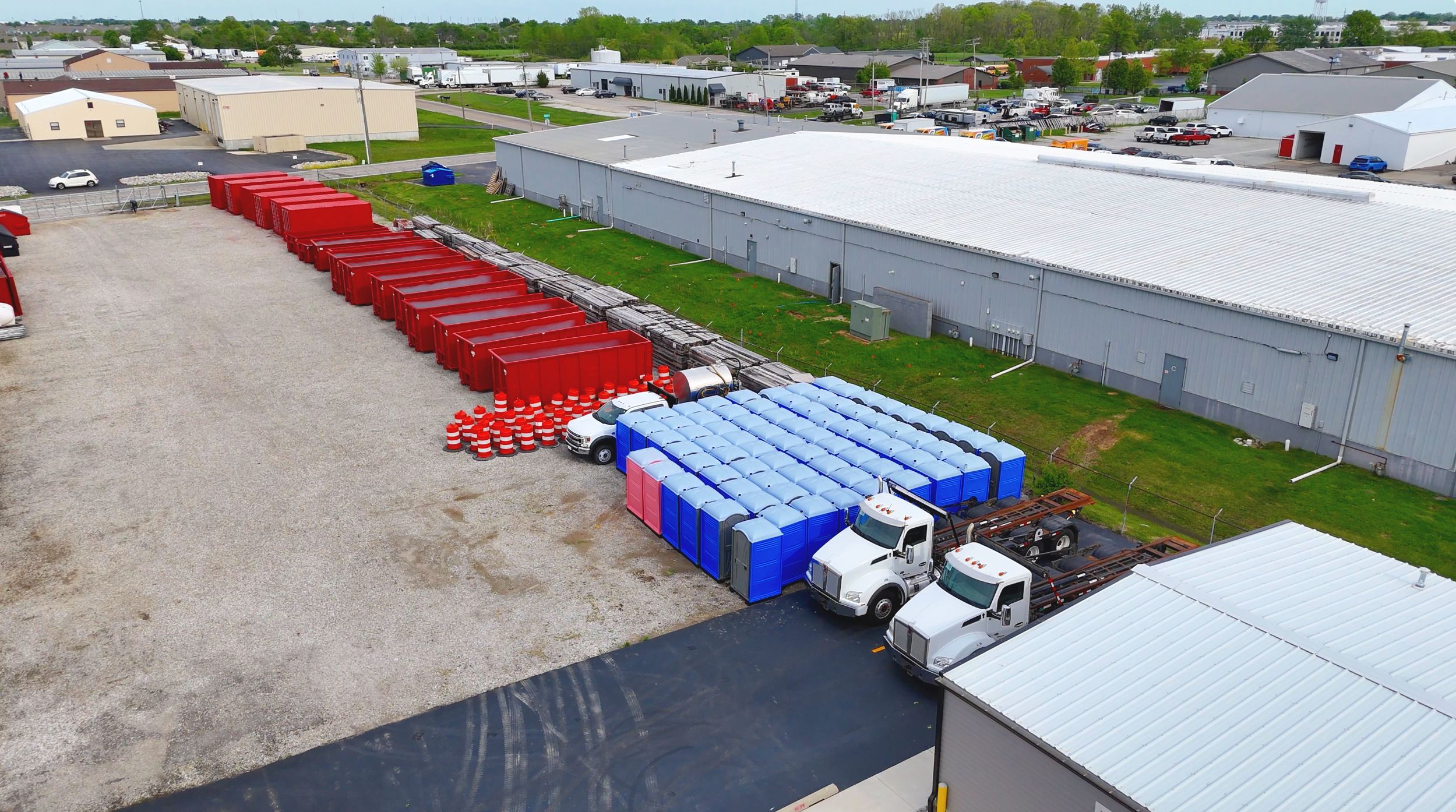 Aerial view of a building and parking lot with multiple trucks, red dumpsters, and portable toilets, surrounded by other industrial and commercial buildings with roads and greenery in the background.